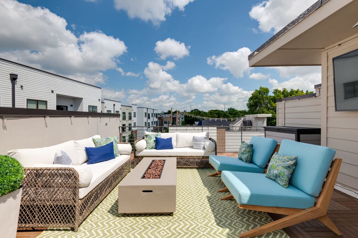 A vibrant rooftop deck features a white wicker sofa with blue and green pillows, two light blue lounge chairs, and a rectangular fire pit on a green and white geometric rug, all under a bright blue sky with white clouds.