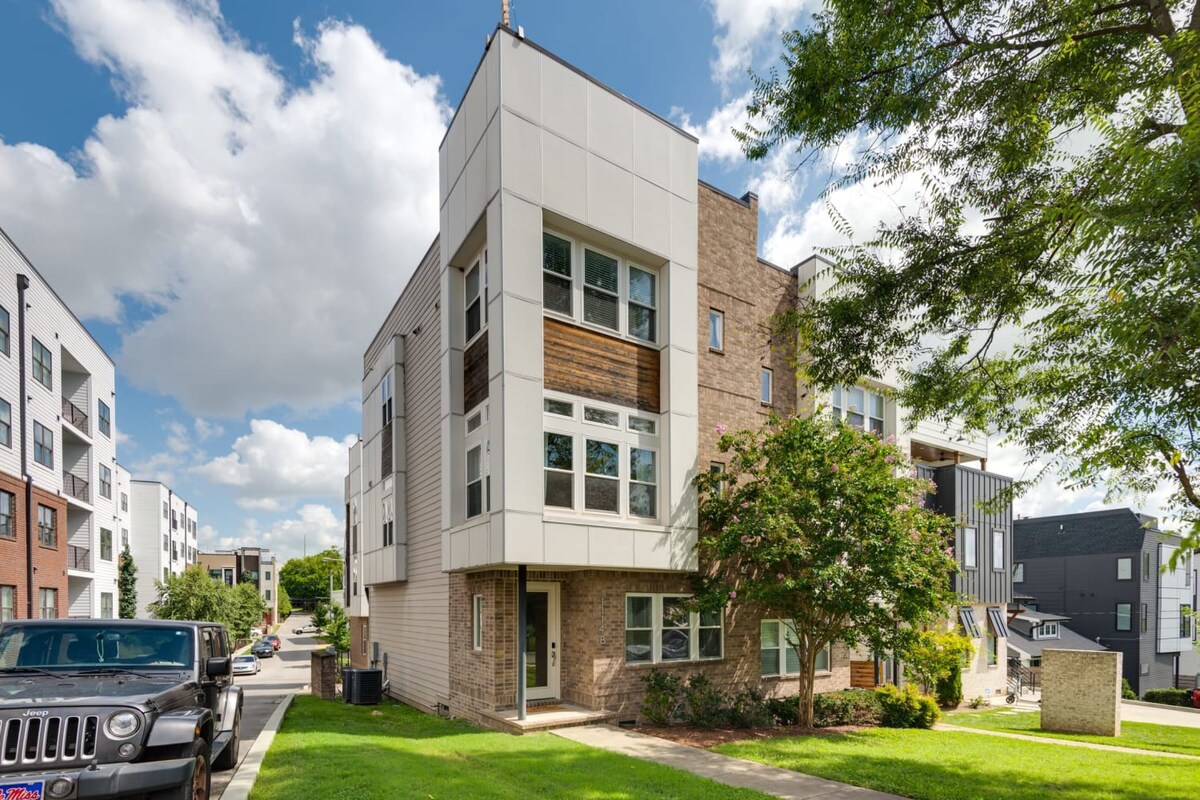 A modern multi-story townhouse features a mix of light brick, white siding, and wood accents, with large windows and a small front yard. A black Jeep is parked on the street in front.