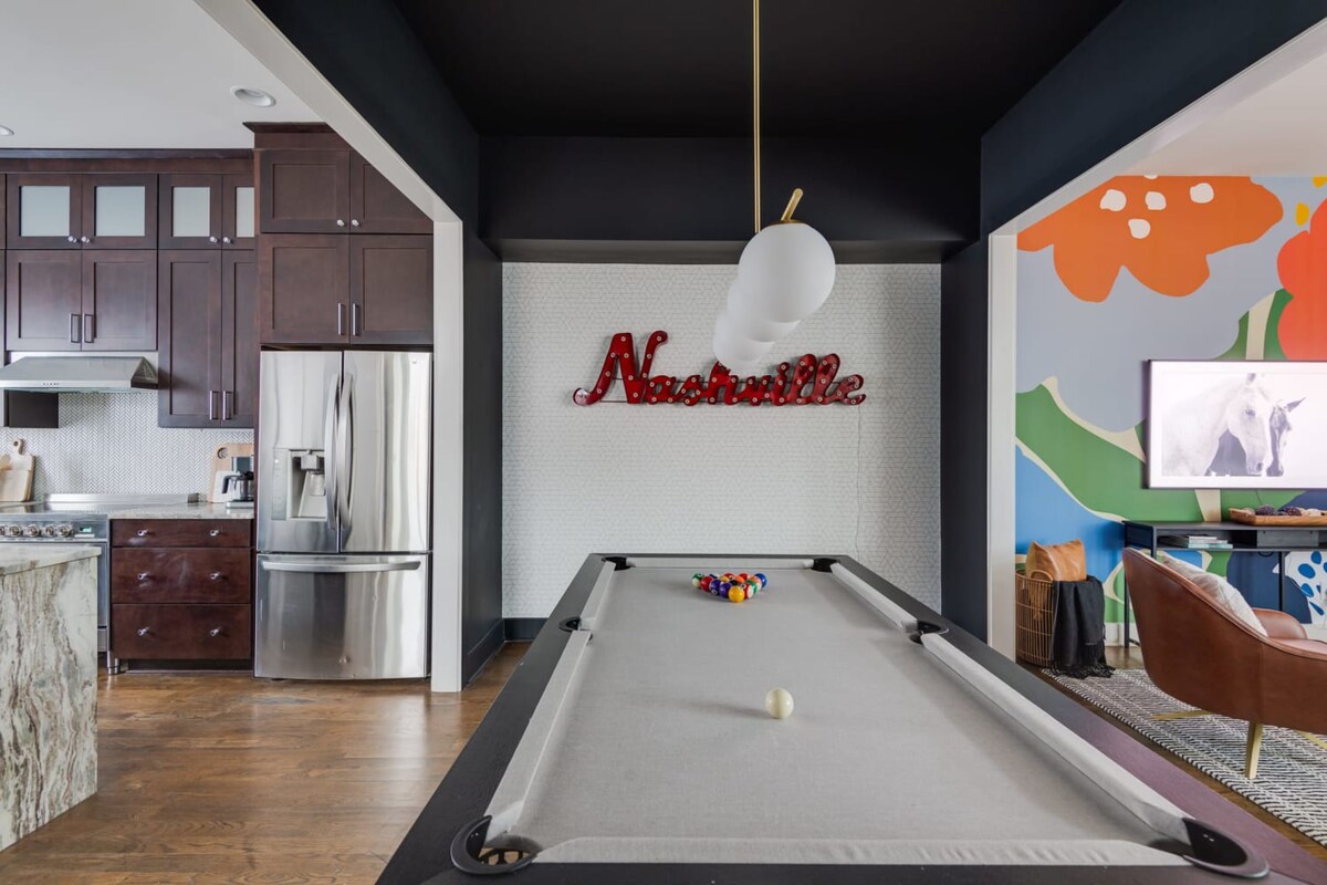 A view of the game room from a different angle, showing the pool table in the foreground and a modern kitchen with dark wood cabinets and stainless steel appliances in the background, connected by an open layout.