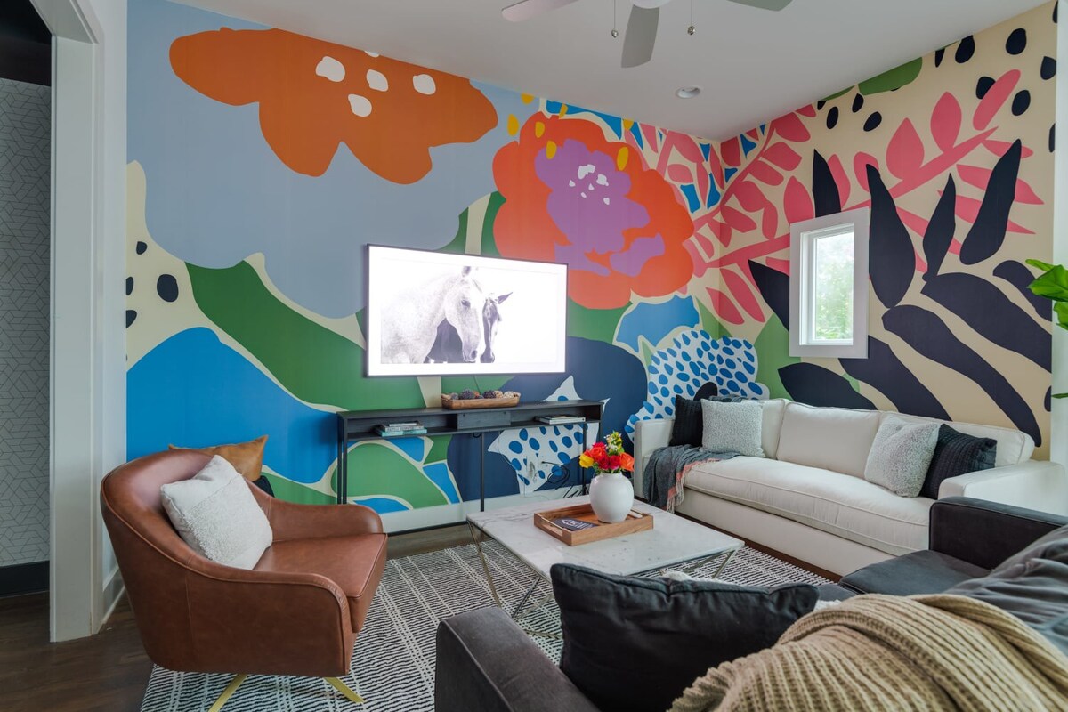 A vibrant living room features a large, colorful abstract mural covering two walls, a white sofa, a brown armchair, and a flat-screen TV on a black console, all centered around a marble coffee table on a patterned rug.