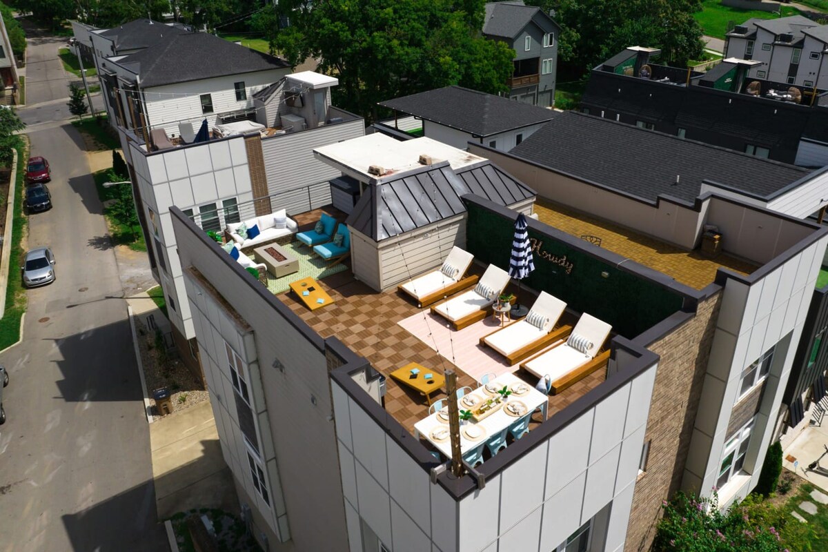 A slightly different aerial perspective of the modern townhouse, showcasing the expansive rooftop deck with multiple seating areas, sun loungers, and a dining table, set against a backdrop of other homes and greenery.