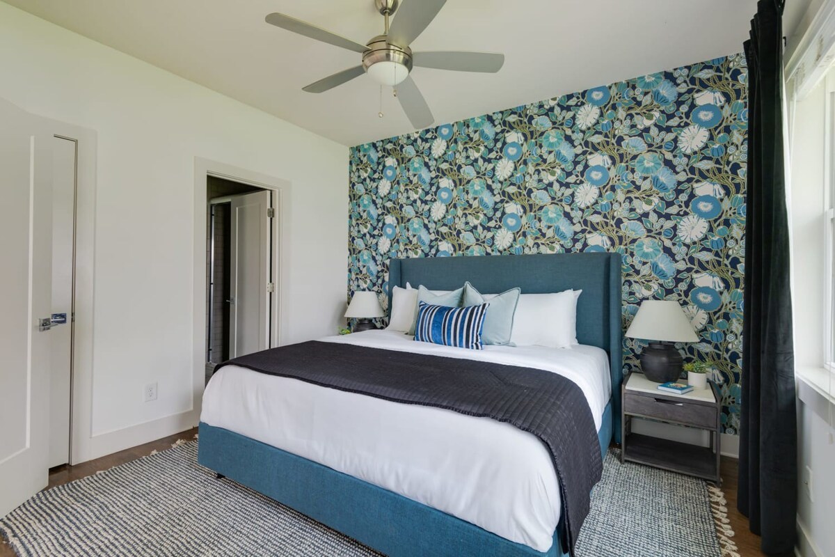 A bright bedroom features a blue upholstered bed with white and blue bedding, a striking blue and green floral wallpaper accent wall behind the bed, and a ceiling fan, with dark curtains framing a window.