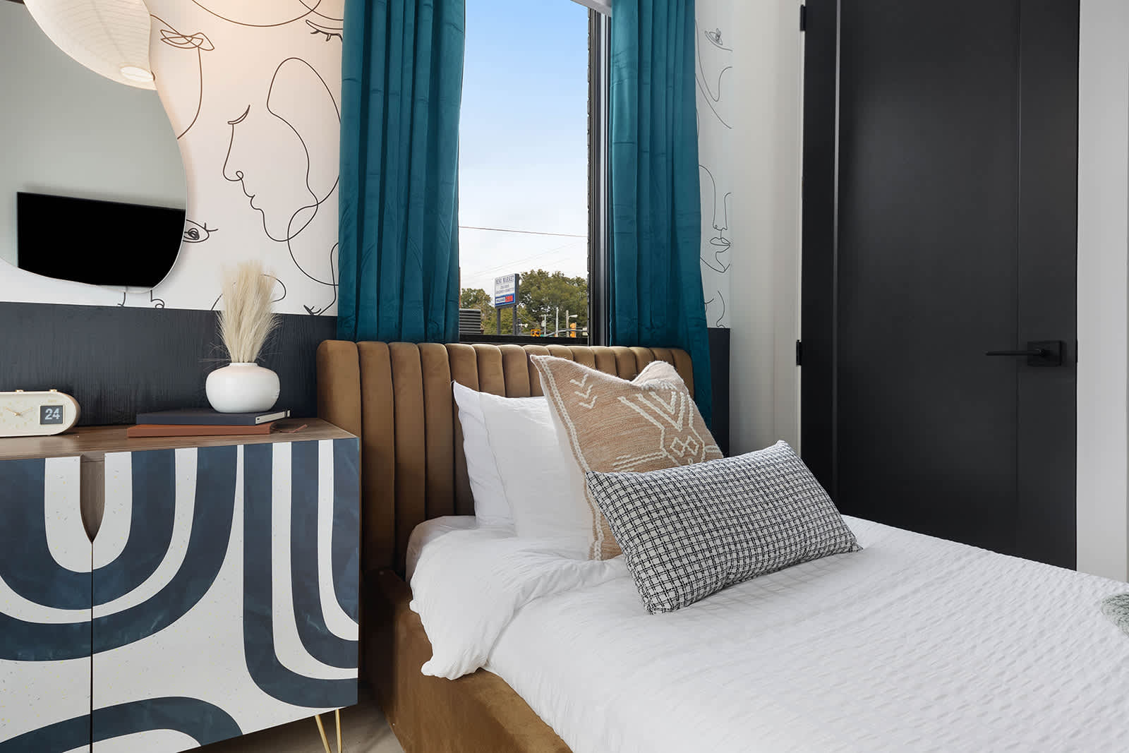 Close-up of one twin bed in a vibrant bedroom, featuring a brown velvet headboard, white bedding, and decorative pillows. A patterned cabinet with gold legs serves as a nightstand, set against a wall with an abstract face mural.