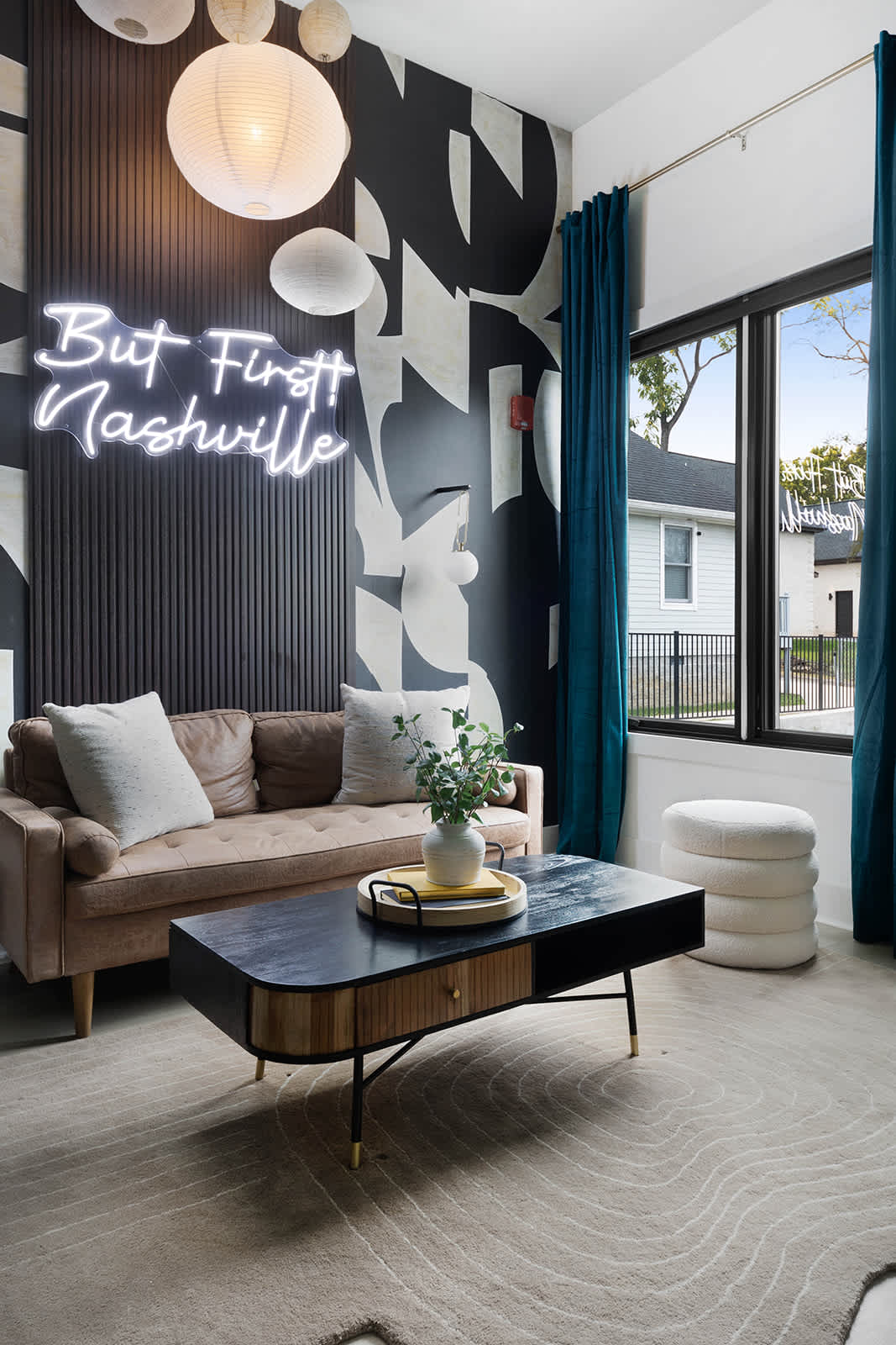 Modern living room featuring a brown velvet sofa, a black and wood coffee table, and a striking "But First Nashville" neon sign against a black and white abstract wall mural. Teal curtains frame a large window, complementing the contemporary design.