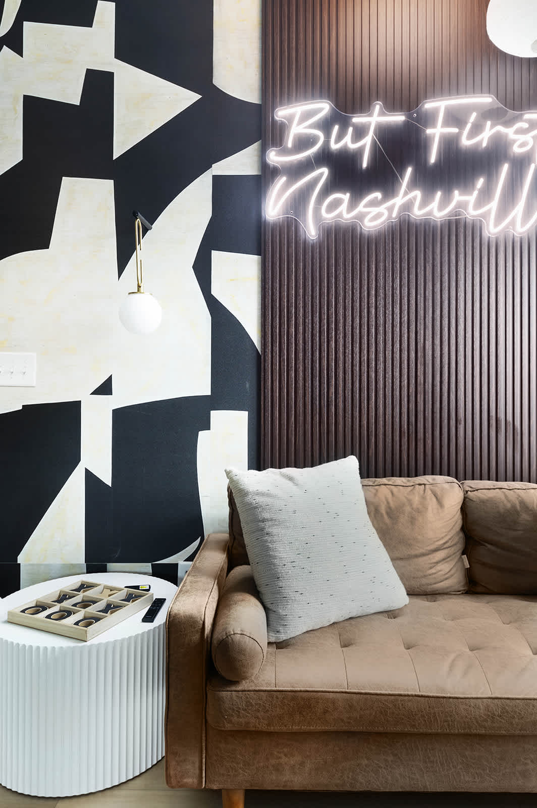 Cozy corner of a living room with a brown velvet sofa, a white fluted side table holding a Tic-Tac-Toe game and other board games. A black and white abstract mural covers the wall, adjacent to a dark wood slatted panel.