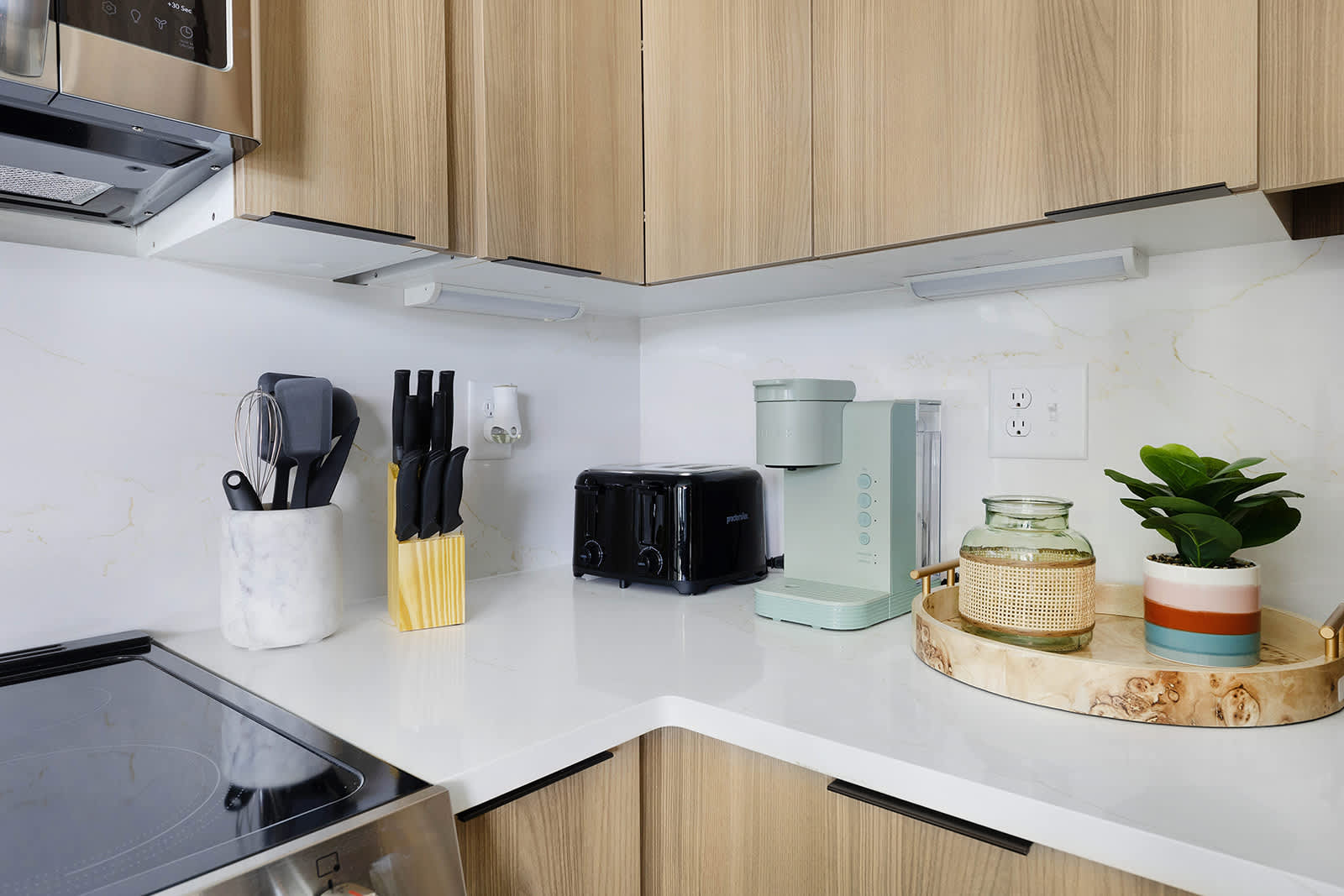 Bright kitchen counter space with white quartz backsplash, light wood upper cabinets, featuring a light green coffee maker, black toaster, and various kitchen utensils in a marble holder.