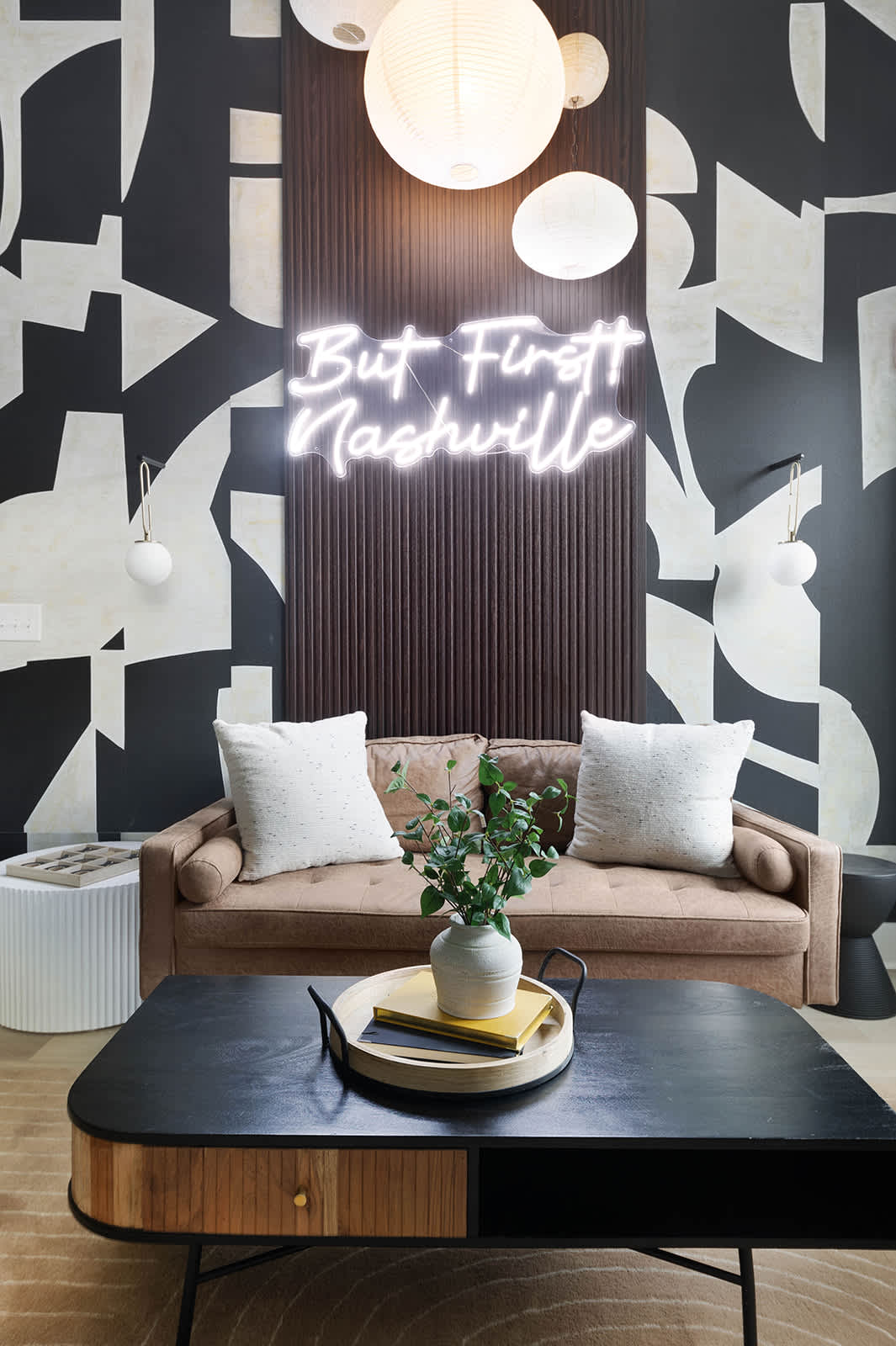 Stylish living room detail showing a brown sofa with white throw pillows, a black and wood coffee table with a decorative tray, and a prominent "But First Nashville" neon sign against a dark wood slatted panel.