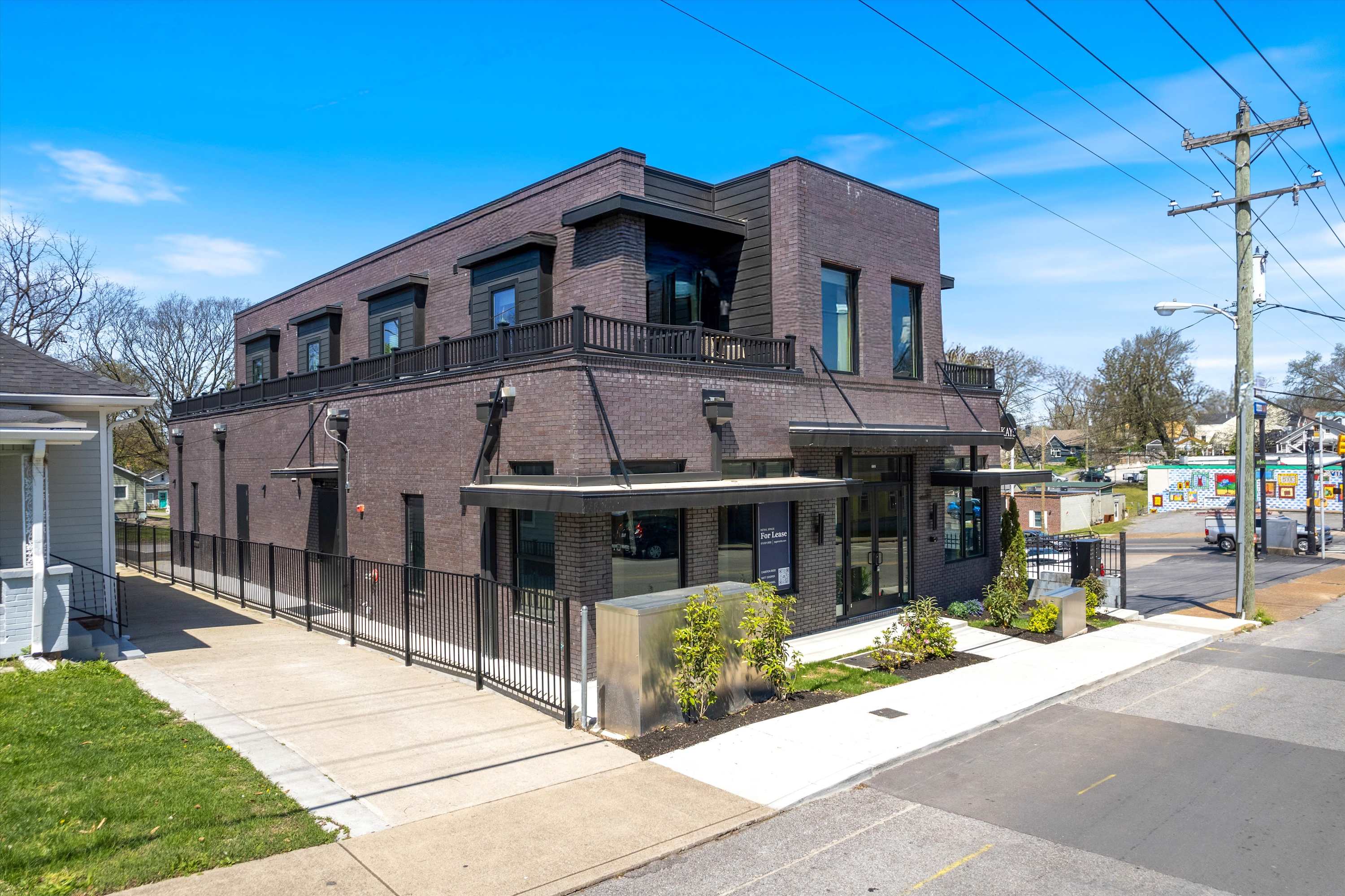A wide aerial shot showcasing the modern brick building with dark accents and multiple windows, featuring a fenced side yard and a paved driveway leading to the rear. The property is surrounded by mature trees and residential homes.