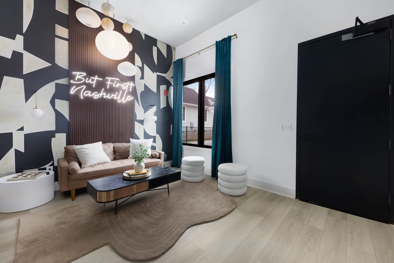 Spacious living room with a brown sofa, black and wood coffee table, and a large black and white abstract mural with a "But First Nashville" neon sign, featuring teal curtains and a wavy area rug.