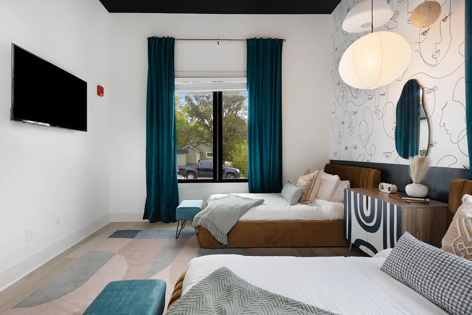 Bright bedroom with two twin beds, a colorful abstract rug, and a wall-mounted TV. Teal curtains frame a large window, and an abstract face mural adds a unique touch to the wall behind the beds.