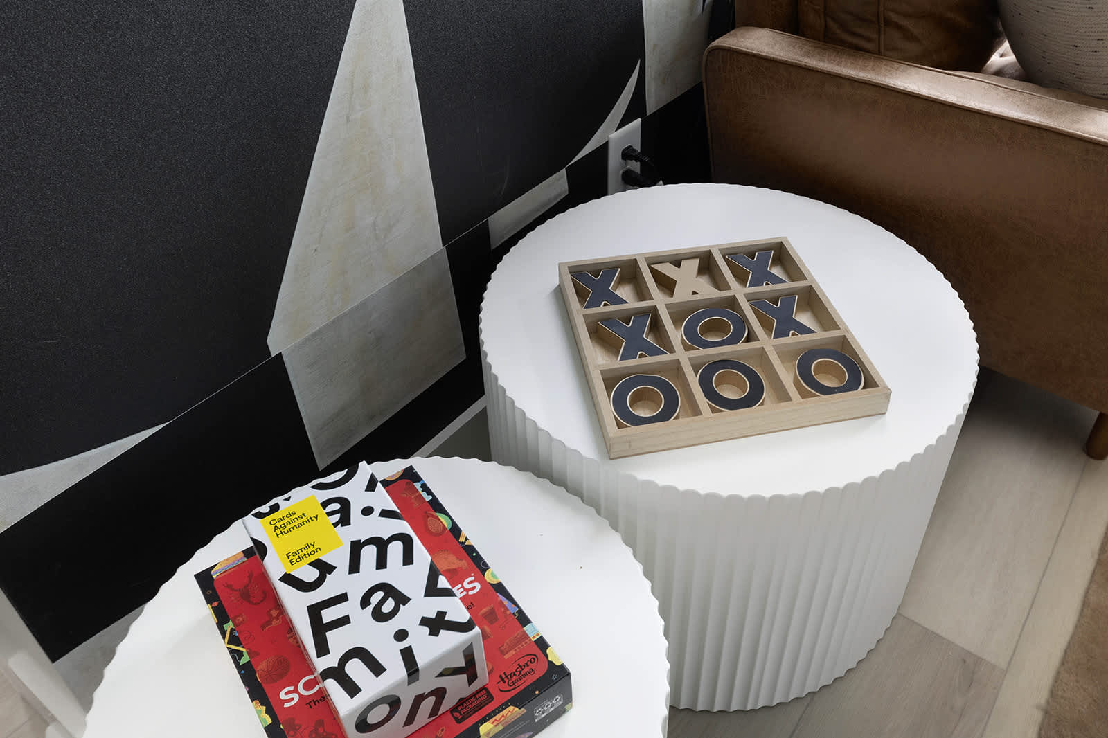 Close-up of two white fluted side tables in the living room, displaying a wooden Tic-Tac-Toe game and various board games. The background features a black and white abstract wall mural, adding a playful and artistic touch.