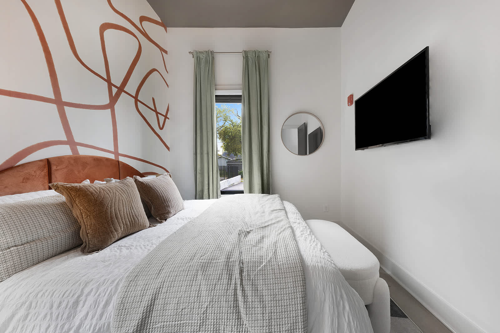 Bright bedroom with a queen-sized bed featuring a rust-colored velvet headboard, white bedding, and a wall-mounted TV. An abstract rust-colored mural adorns one wall, complemented by light green curtains and a round mirror.