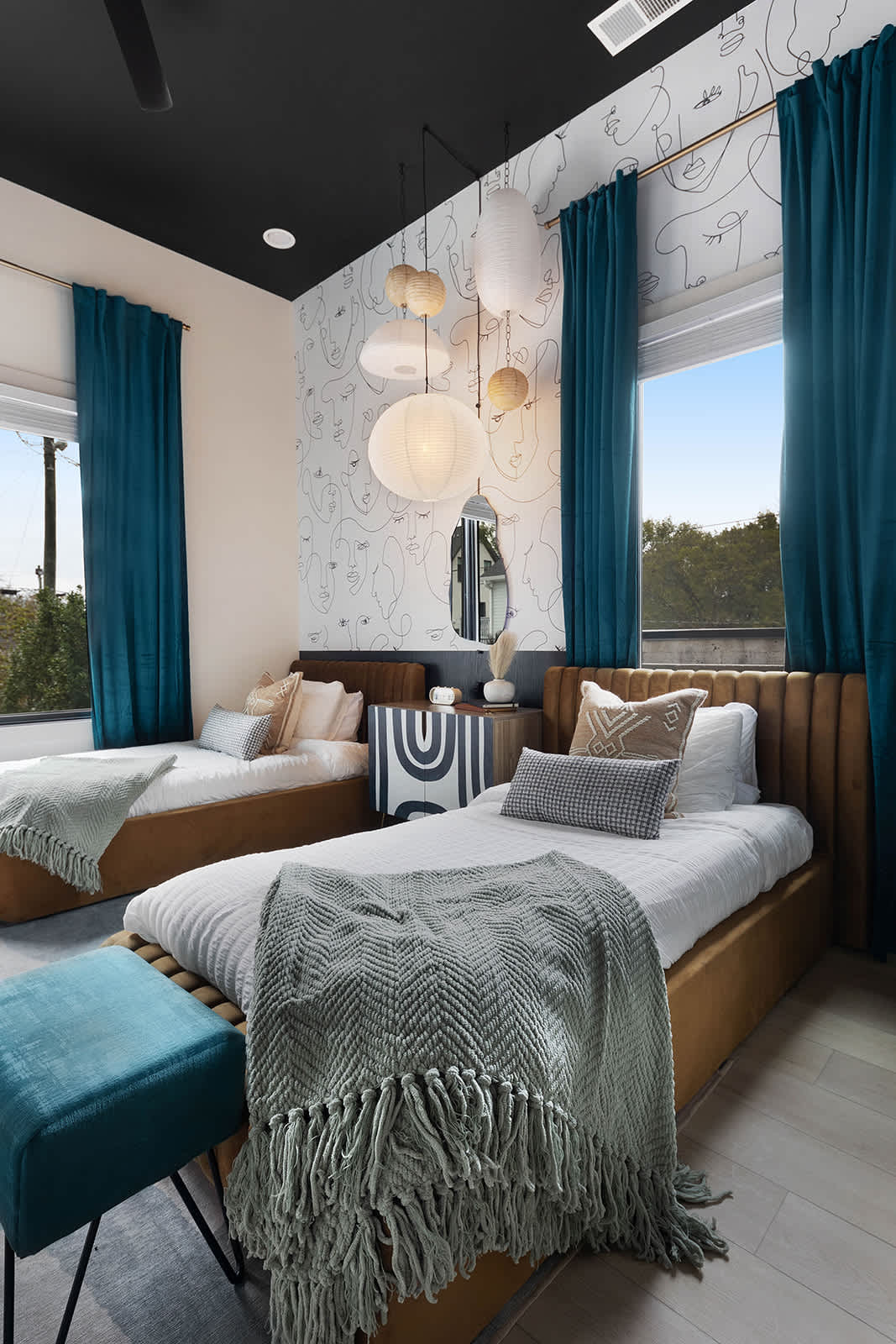 Modern bedroom with two twin beds featuring brown velvet headboards, a shared patterned nightstand, and a black ceiling. An abstract face mural covers the wall, and teal curtains frame two large windows, with decorative paper lanterns overhead.