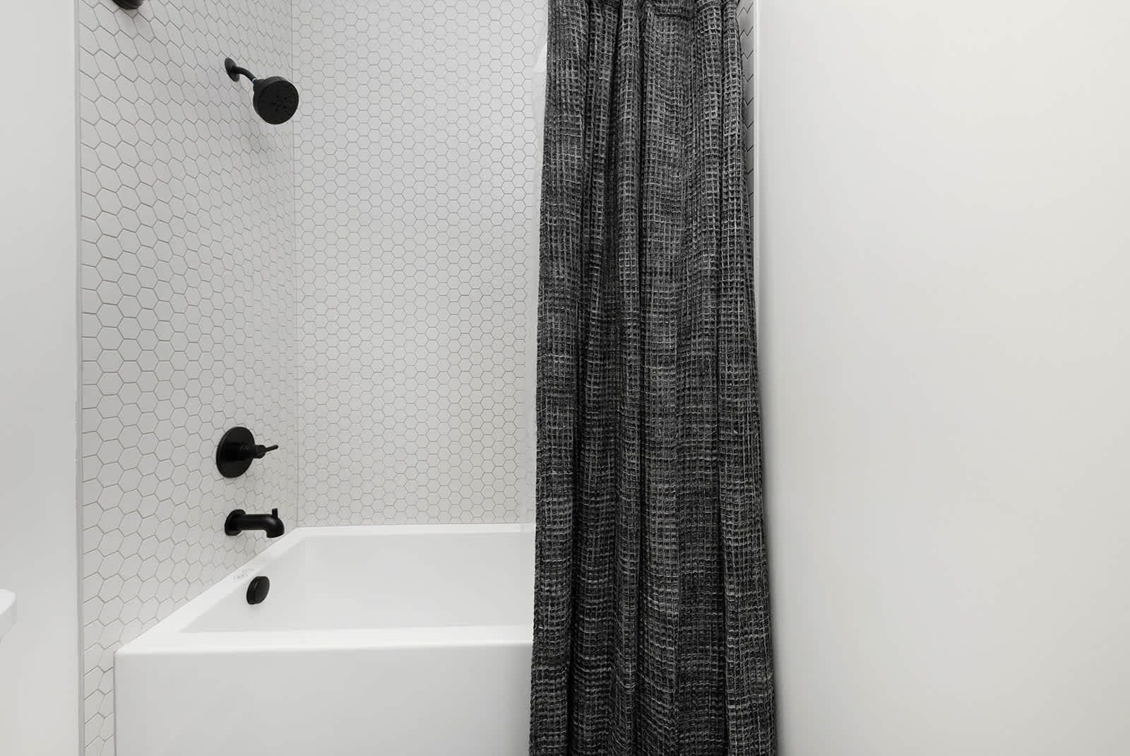 A modern bathroom features a white bathtub with black fixtures, surrounded by white hexagonal tile walls. A dark gray patterned shower curtain hangs beside the tub, complementing the black hardware.