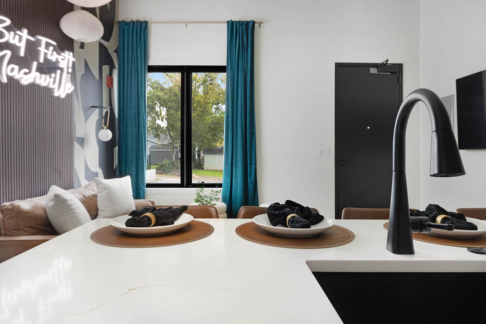 Close-up of a white quartz kitchen island with a black gooseneck faucet and sink, set with place settings, offering a glimpse of the adjacent living area with teal curtains and a neon sign.