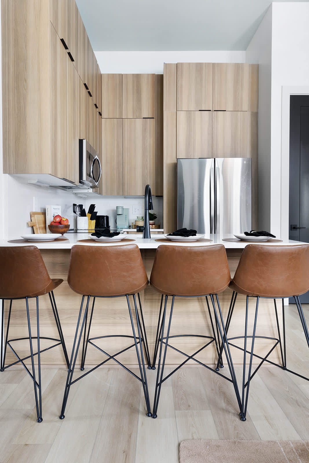 Full view of the modern kitchen featuring light wood cabinetry, stainless steel refrigerator, and a white quartz island with four brown leather bar stools, set against light wood flooring.