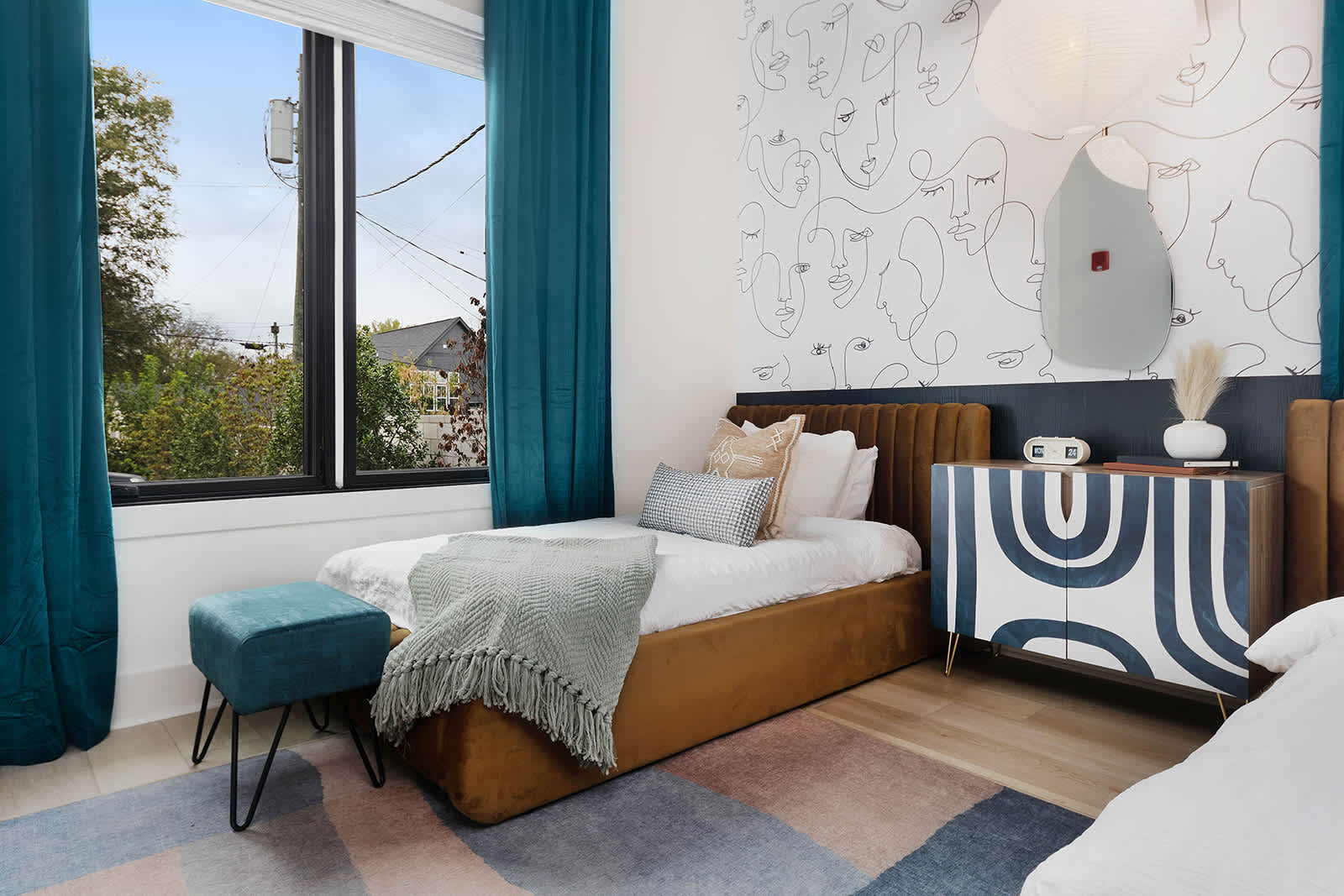 Vibrant bedroom featuring two twin beds with brown velvet headboards, a colorful abstract rug, and a unique wall mural depicting abstract faces. Teal curtains frame a large window, and a patterned cabinet sits between the beds.