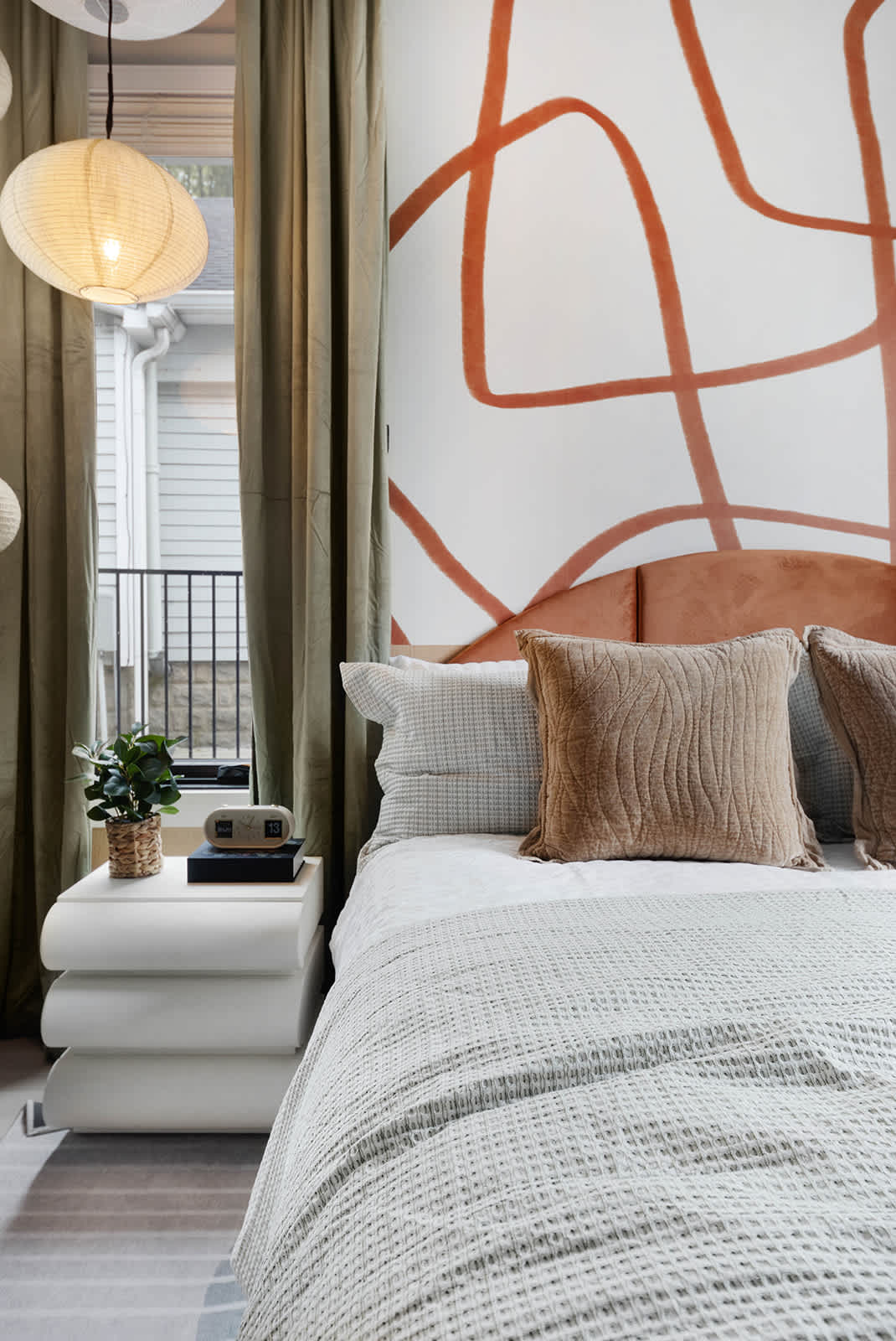 Close-up of a bedroom showing a bed with a rust-colored velvet headboard, white and textured bedding, and a white stacked nightstand with a small plant and alarm clock. Light green curtains frame a window, with decorative paper lanterns hanging nearby.