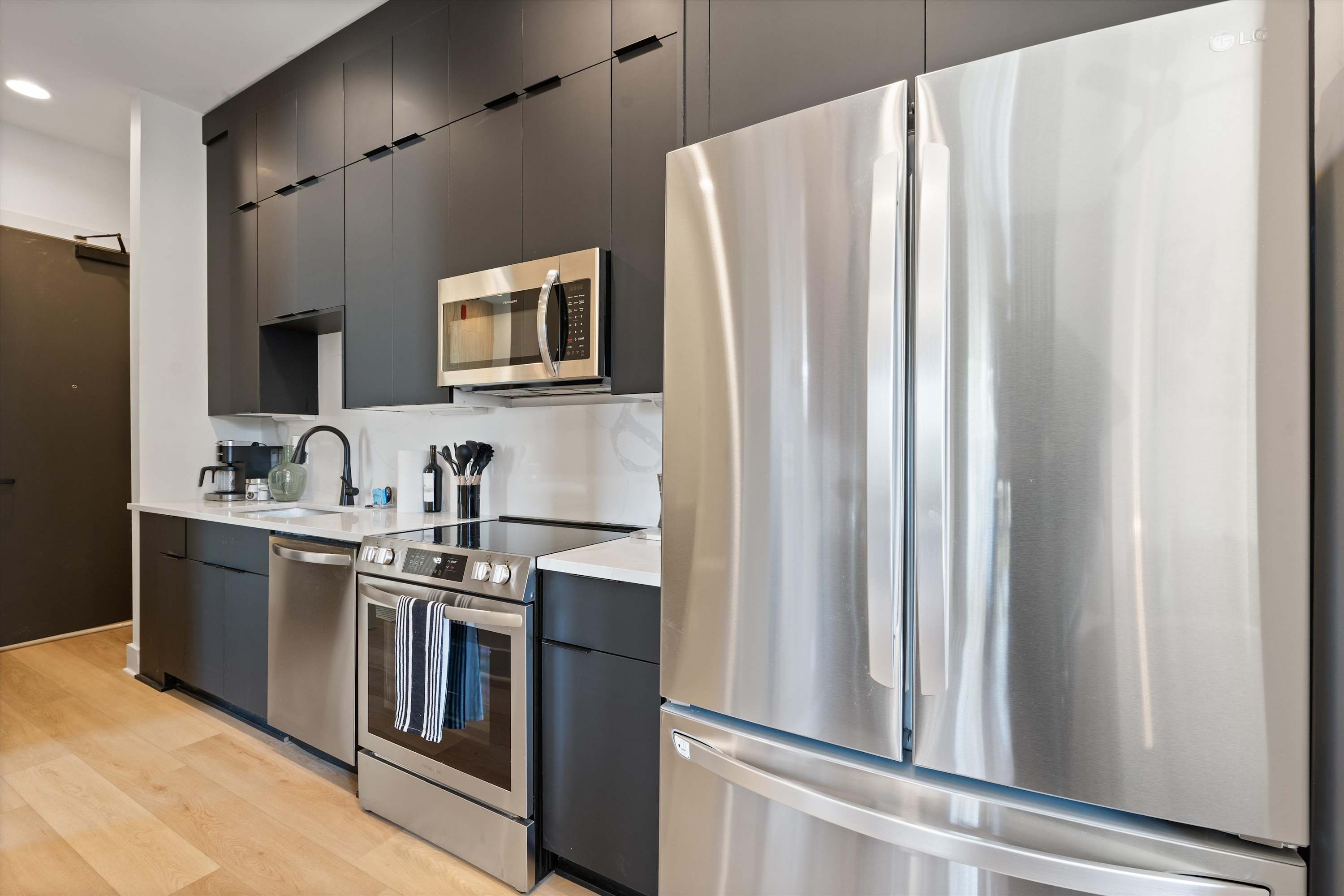 This view of the kitchen emphasizes the full suite of stainless steel appliances, including a large French door refrigerator, dishwasher, and electric range. Dark cabinetry and white countertops maintain the sleek, contemporary aesthetic of the space.