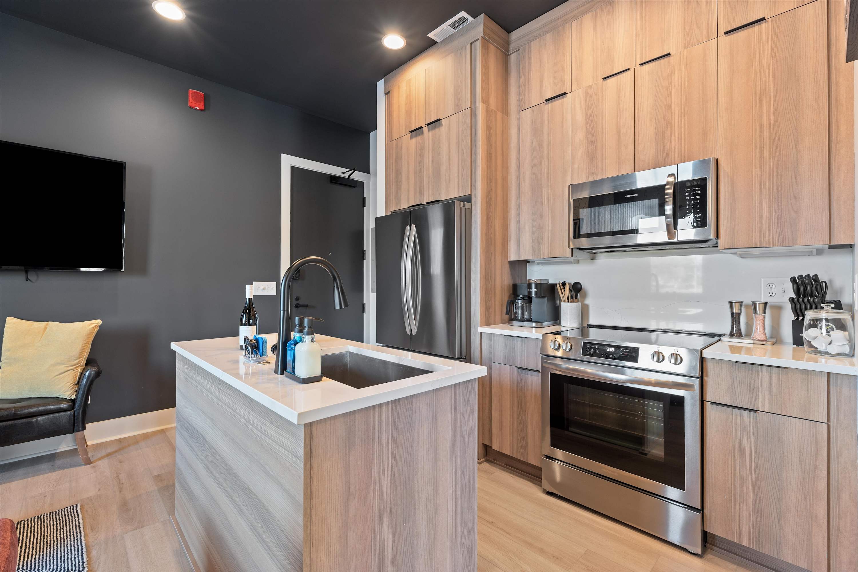 Modern kitchen with light wood cabinetry, stainless steel appliances including a refrigerator, microwave, and electric range. A central island features an undermount sink, complemented by dark gray walls and light hardwood floors.
