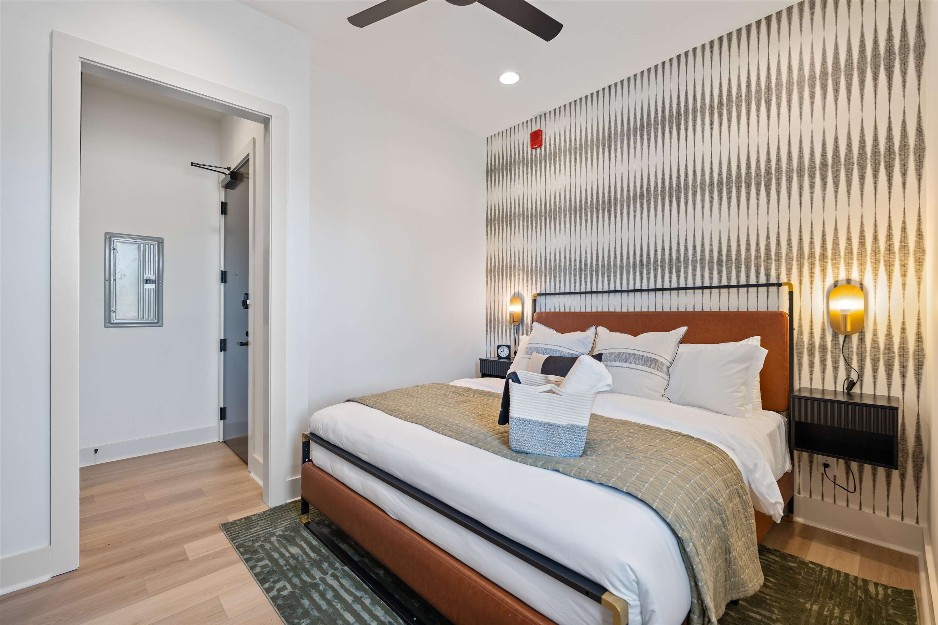 Stylish bedroom with a queen-sized bed featuring a brown leather headboard and a patterned throw blanket, set against a geometric patterned accent wall. Two wall-mounted nightstands with lamps flank the bed.