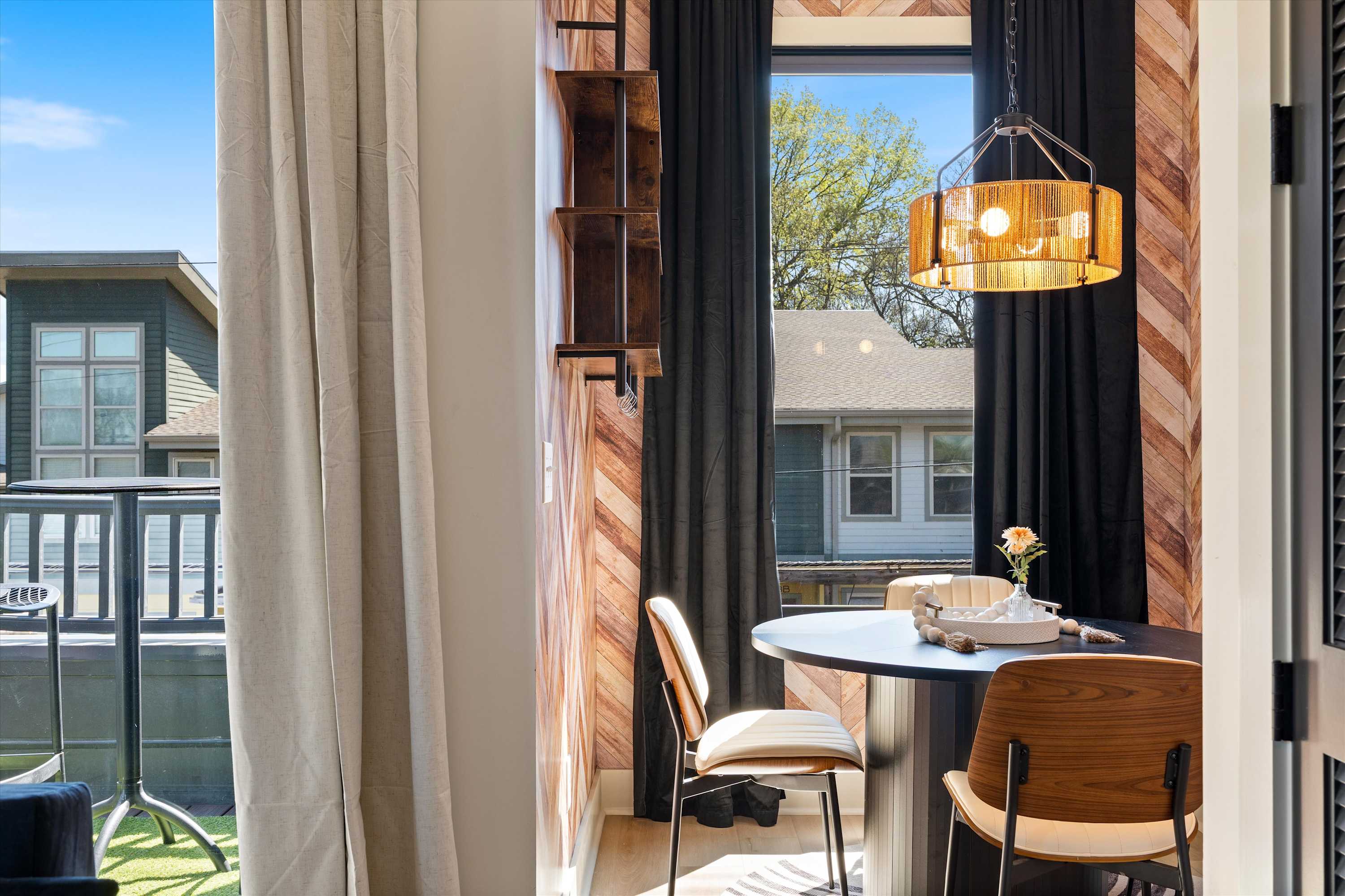 Dining area with a round table and chairs, featuring a large window with both sheer and dark curtains, offering a glimpse of an outdoor balcony and the neighborhood beyond. A wooden shelf unit is mounted on the wall.