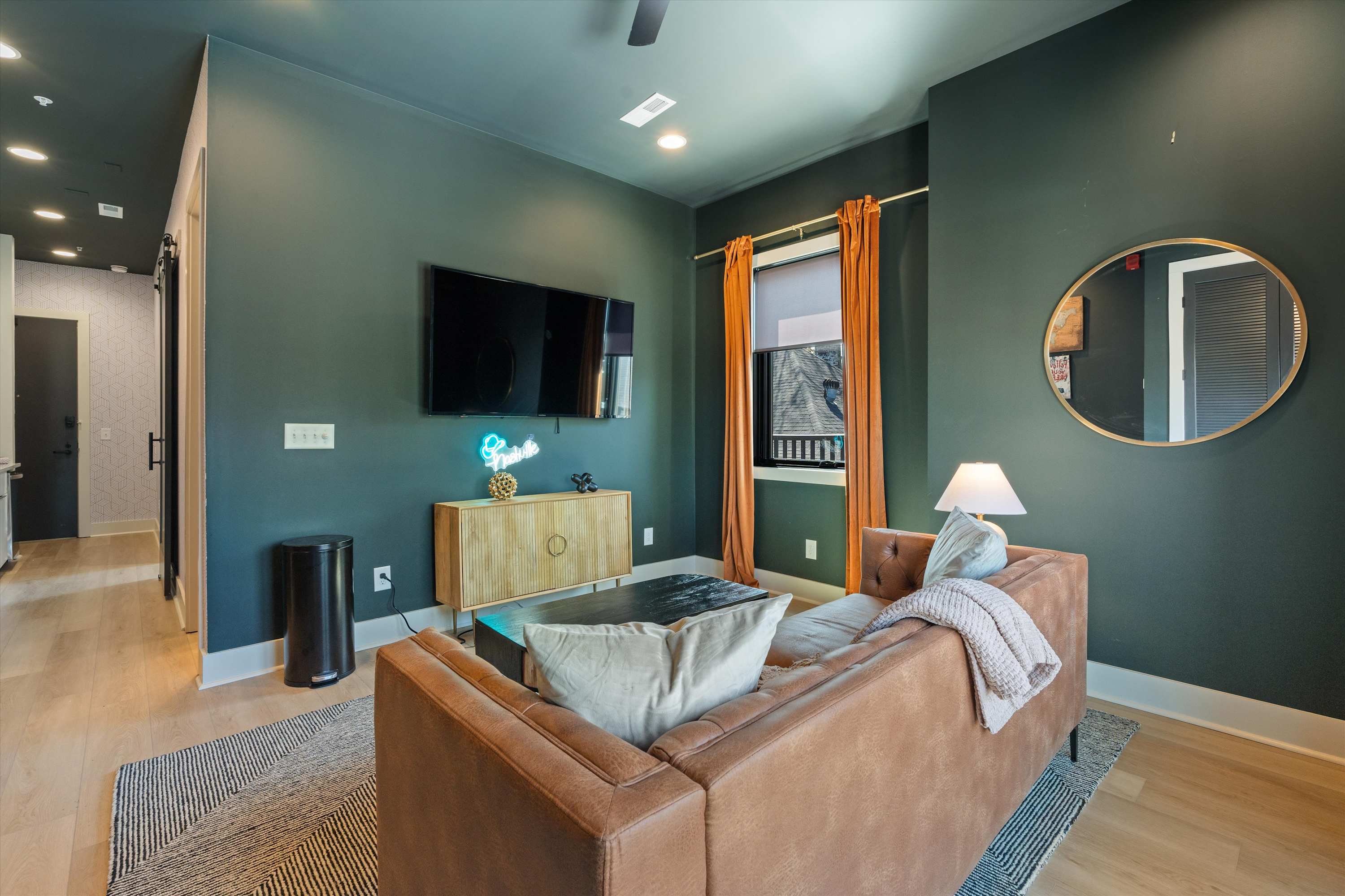 This living room view shows a brown leather sofa with throw pillows, facing a wall-mounted flat-screen TV above a wooden console table with a neon sign. Dark green walls, orange curtains, and a geometric area rug contribute to the room's vibrant and modern aesthetic.