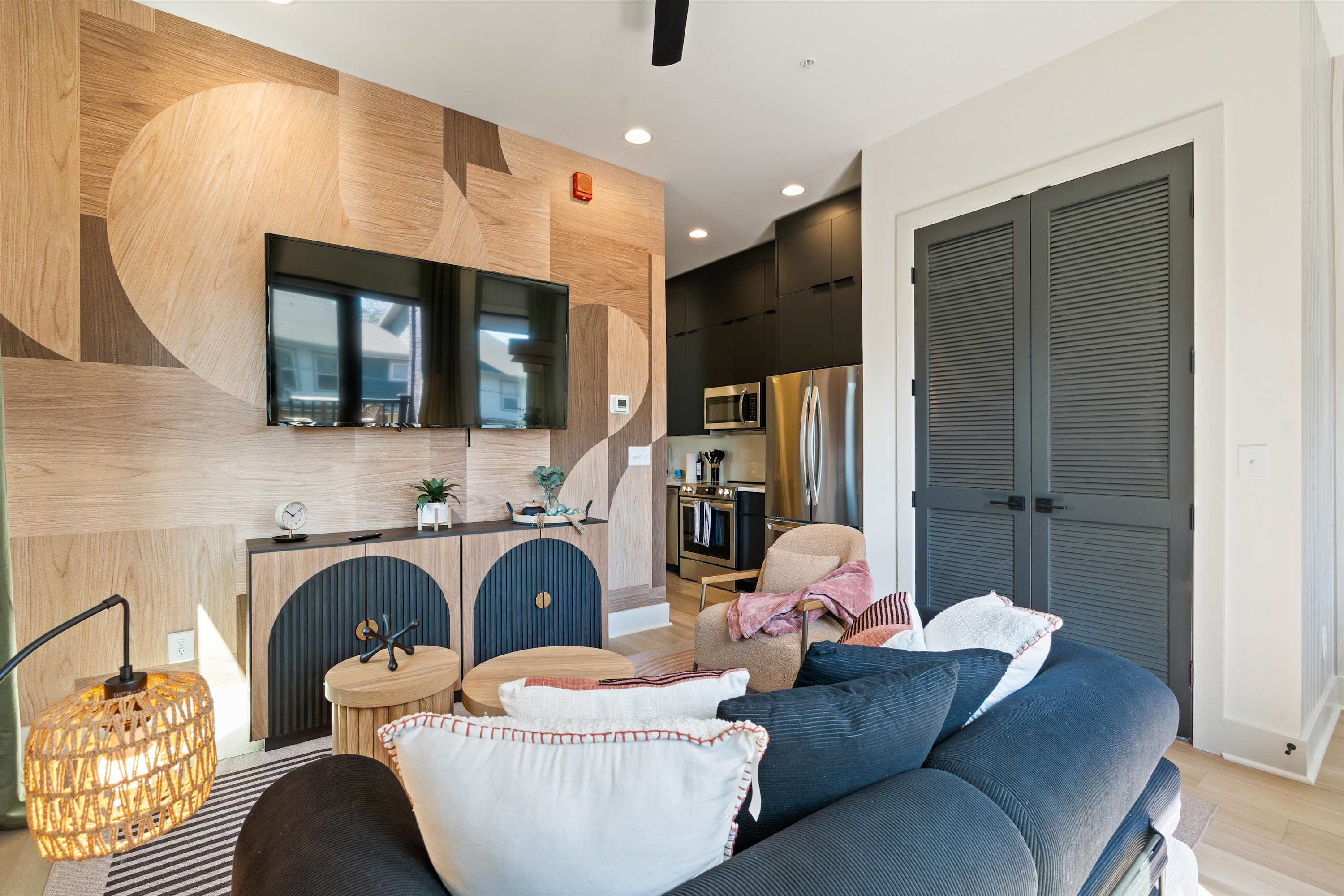 Open concept living space with a dark sofa and a geometric wood panel accent wall with a mounted TV, looking towards a modern kitchen with dark flat-panel cabinets and stainless steel appliances.