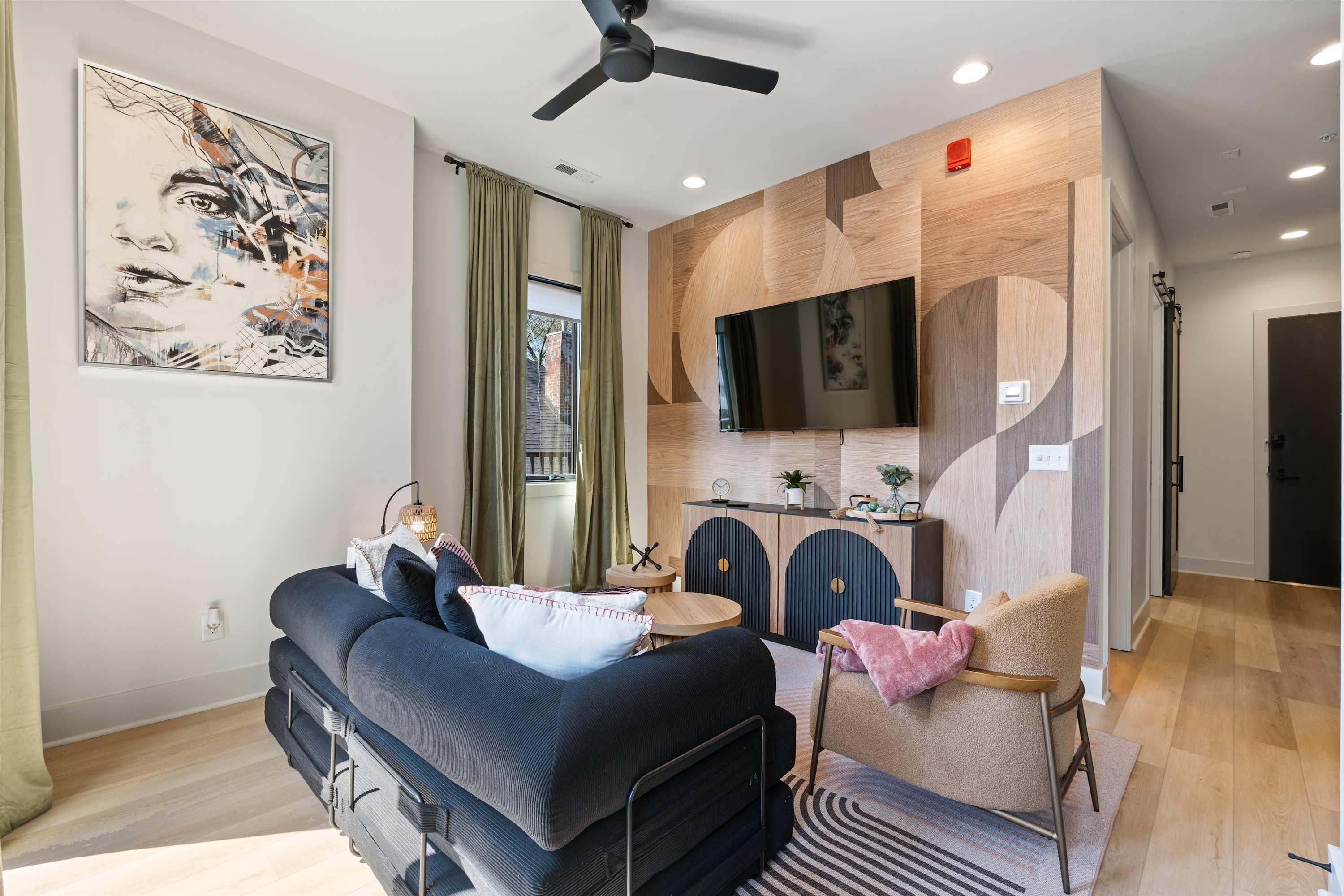 Modern living room featuring a dark sofa, a light-colored armchair, and a wall-mounted TV set against a geometric wood panel accent wall. A ceiling fan and large window with green curtains are also visible.