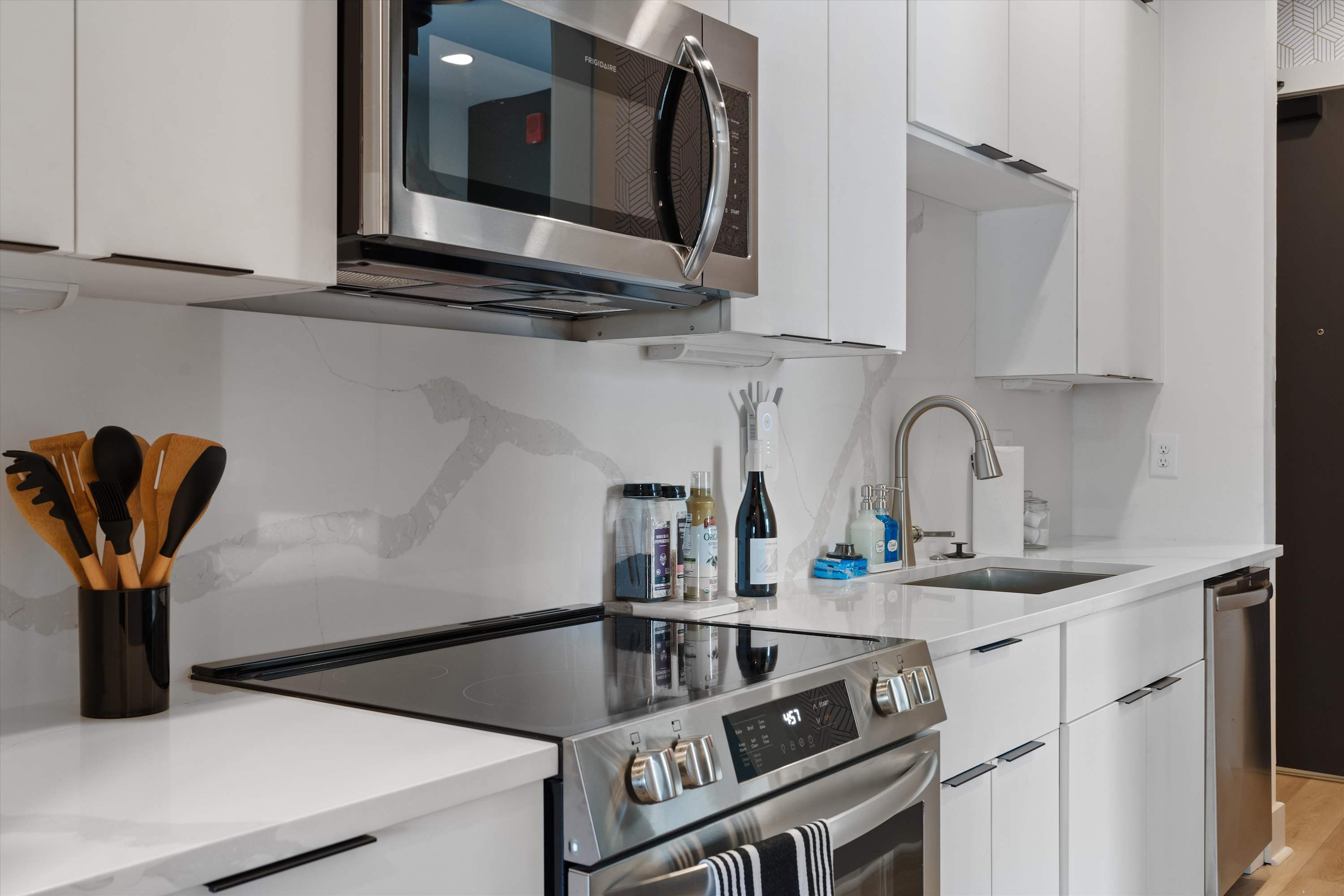 Close-up of a modern kitchen showing a stainless steel microwave above an electric range with a black cooktop. White quartz countertops and a matching veined backsplash are visible, along with a stainless steel sink and faucet.