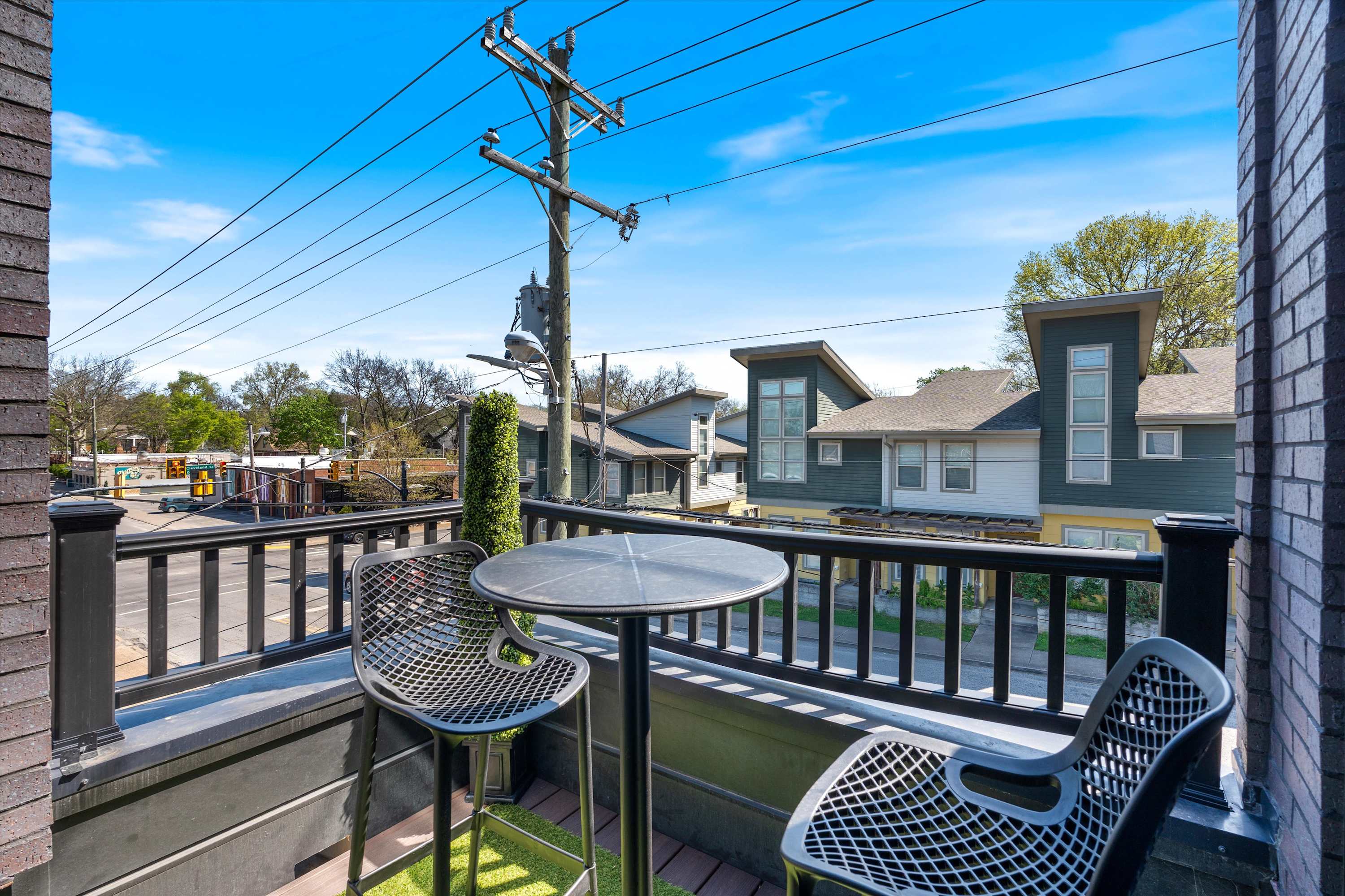A cozy balcony space with a black metal railing, furnished with a round black table and two matching mesh chairs, offering a view of the street and residential area.