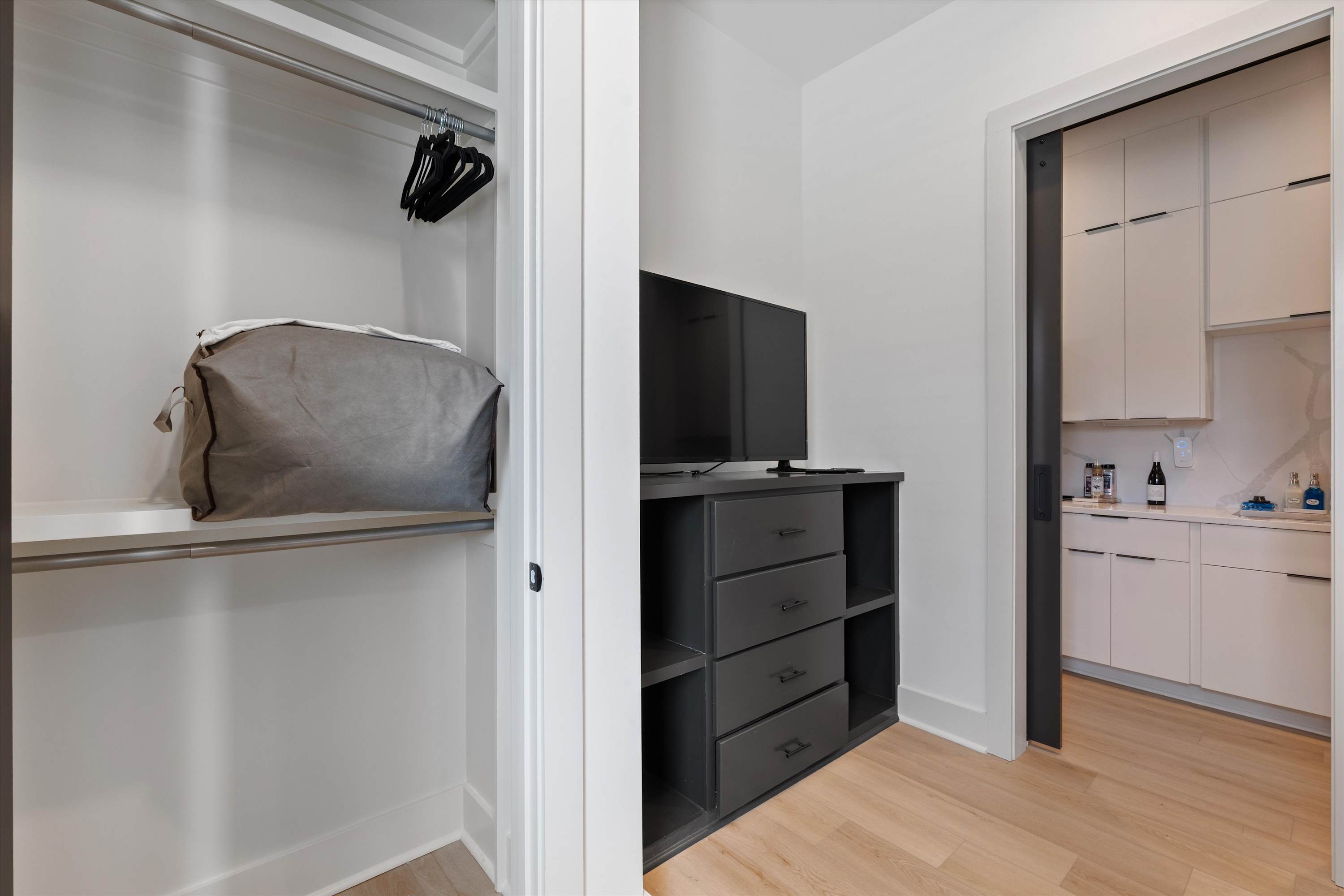 Transitional space featuring a built-in closet with shelving and hangers, and a dark dresser with a television. Light hardwood floors connect to the modern white-cabinet kitchen visible in the background.