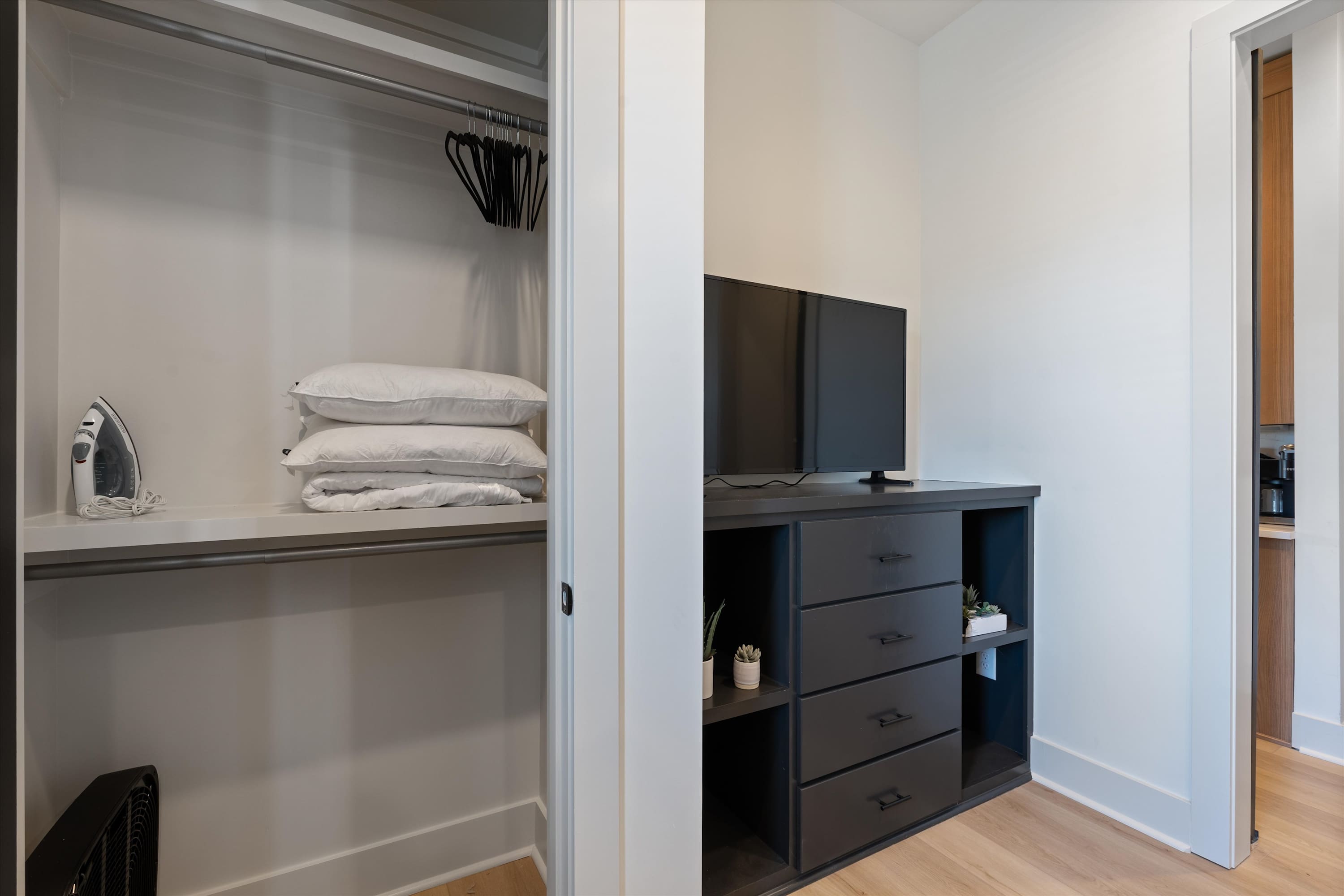 A bedroom closet is neatly organized with hanging space and shelves holding extra pillows and an iron, adjacent to a dark dresser with open shelving and drawers. The light hardwood floors extend throughout the space.