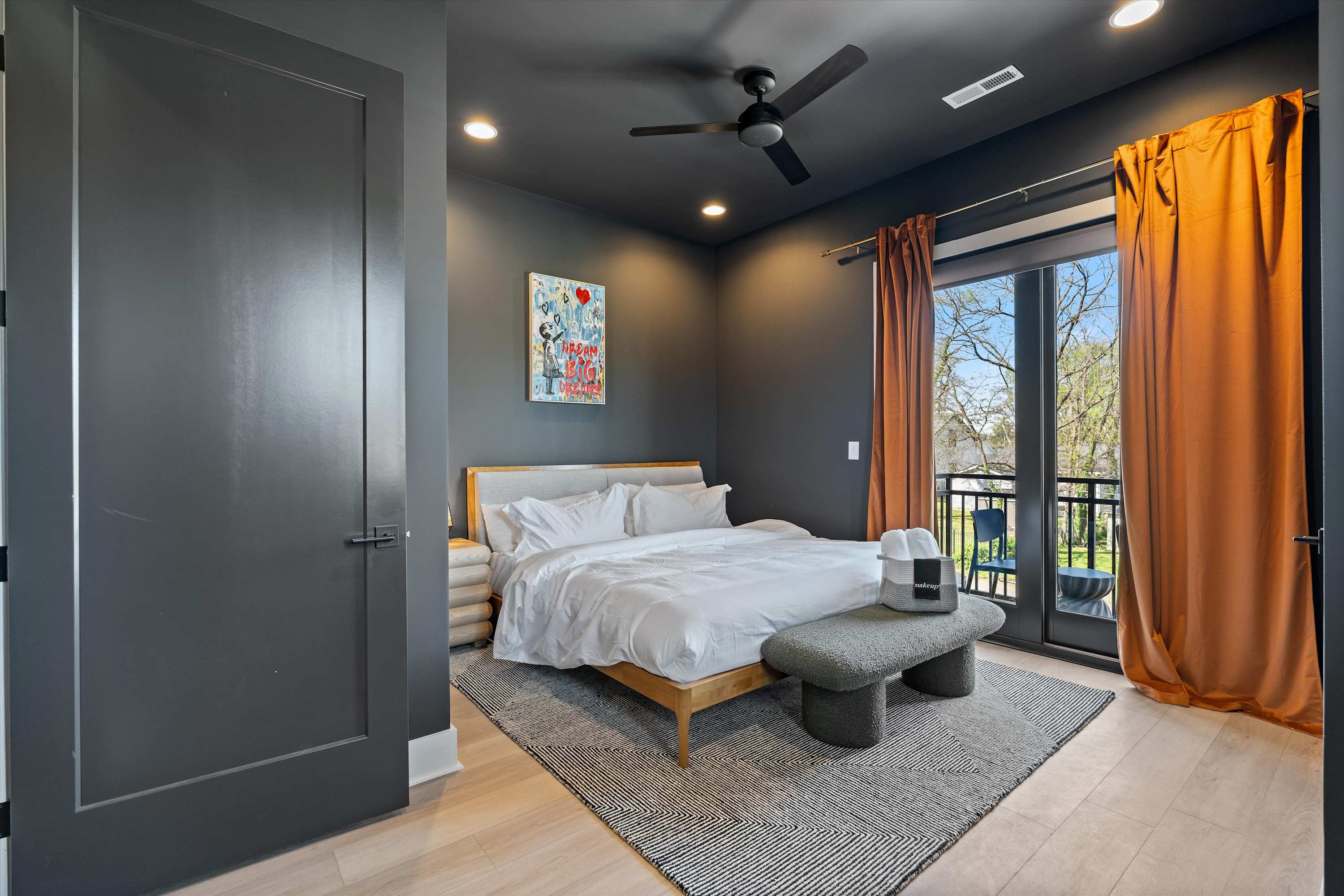 A modern bedroom features a king-sized bed with white bedding and a light wood frame, complemented by a grey textured bench at the foot. Dark grey walls and orange curtains frame a glass door leading to a balcony, with a geometric area rug on light hardwood floors.