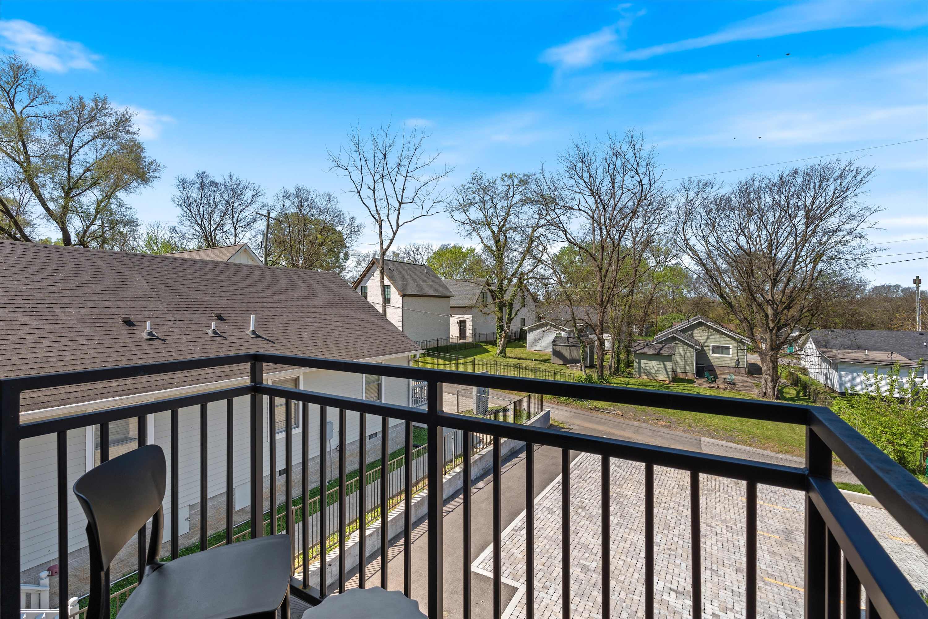 View from a private balcony, showcasing a black metal railing in the foreground and overlooking a paved parking area. Beyond, a residential neighborhood with various house styles and mature trees is visible under a bright blue sky.