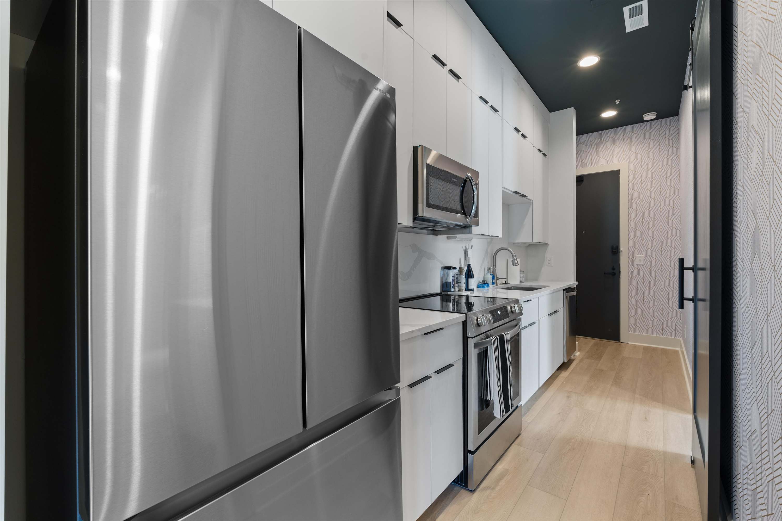 View of a modern galley kitchen with a large stainless steel French door refrigerator on the left, white flat-panel cabinets, and light hardwood flooring. The dark ceiling extends through the space, creating a sleek, contemporary feel.
