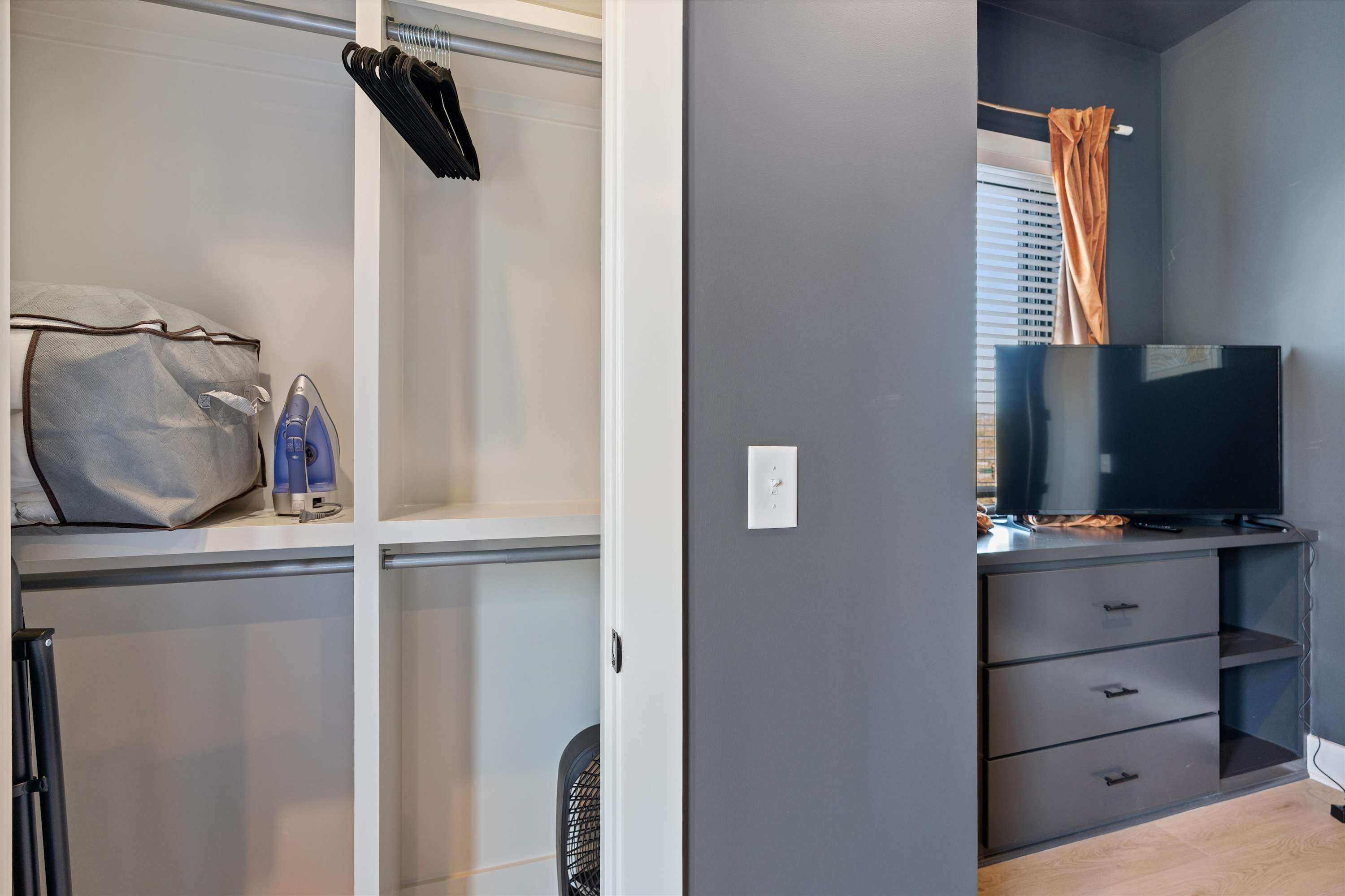 Organized closet space featuring white shelving, hanging rods with black hangers, and an iron. Light hardwood floors are visible, and a dark gray wall with a window is seen to the right, suggesting a bedroom closet.