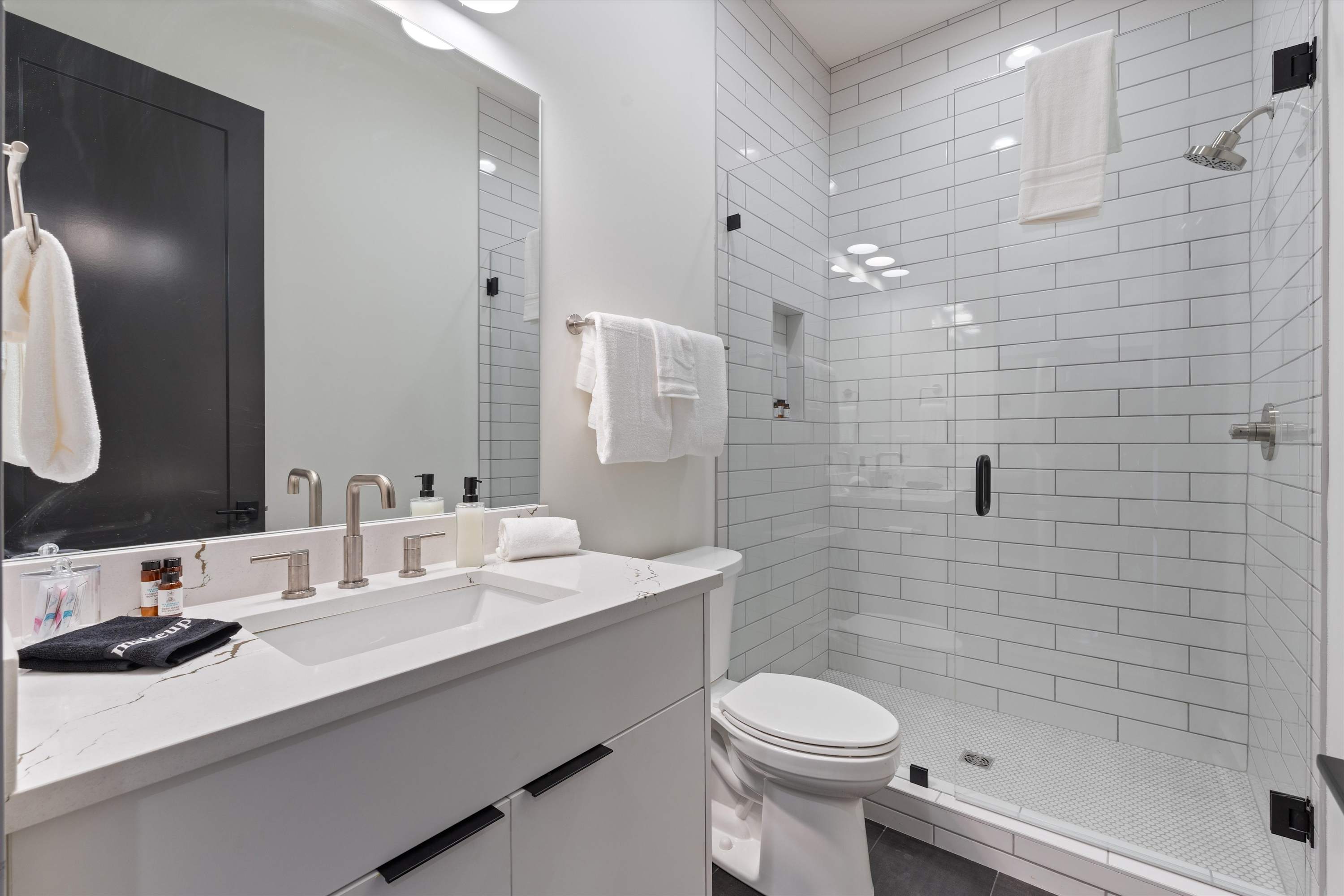 Bright modern bathroom featuring a white vanity with a single sink and brushed nickel faucet, a toilet, and a walk-in shower with white subway tiles and a glass enclosure. Dark floor tiles contrast with the white fixtures.