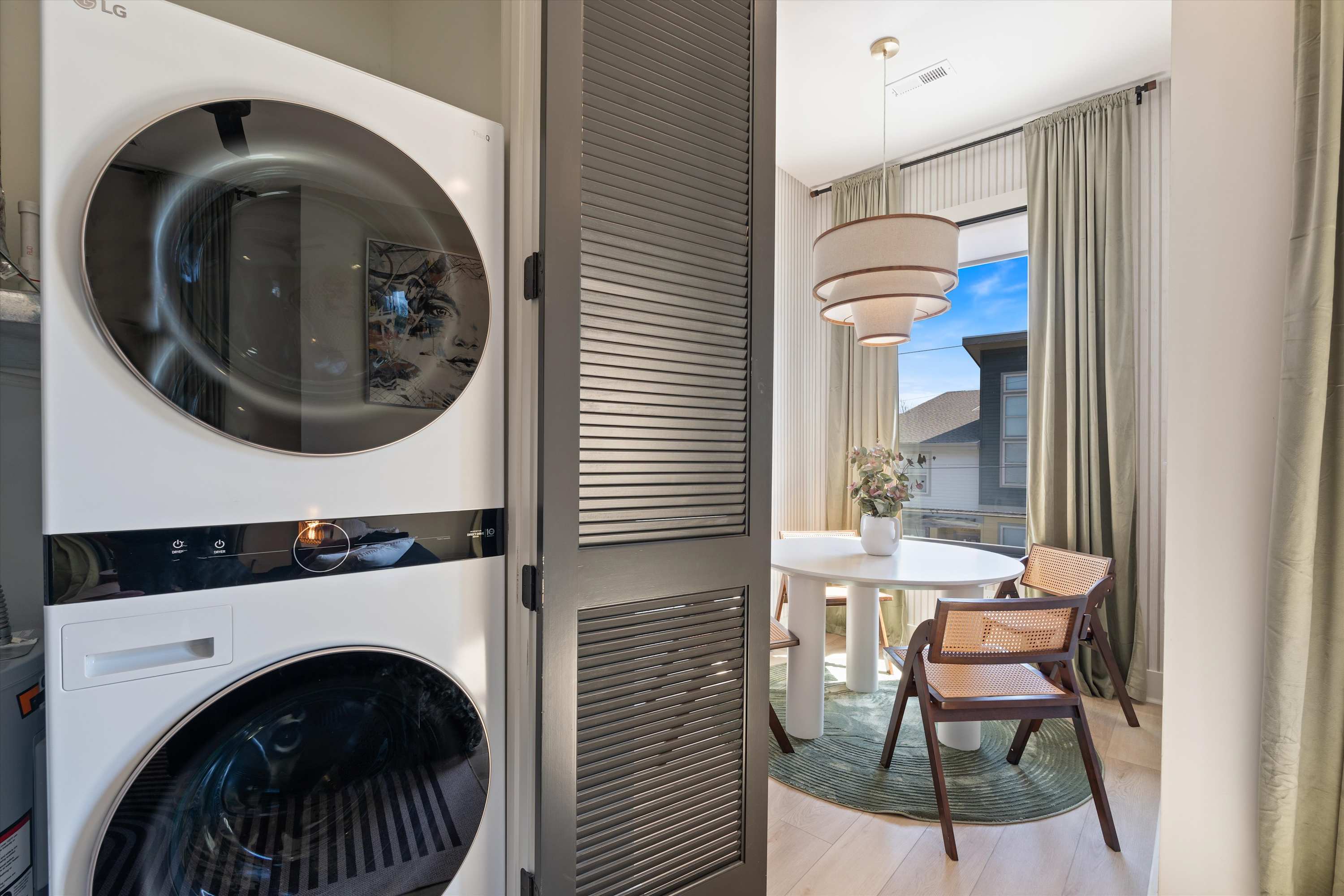Bright dining area featuring a white round table with two modern chairs, set against a large window with light green curtains. A stacked silver washer and dryer unit is visible through an open louvered door, next to a light-colored accent wall.
