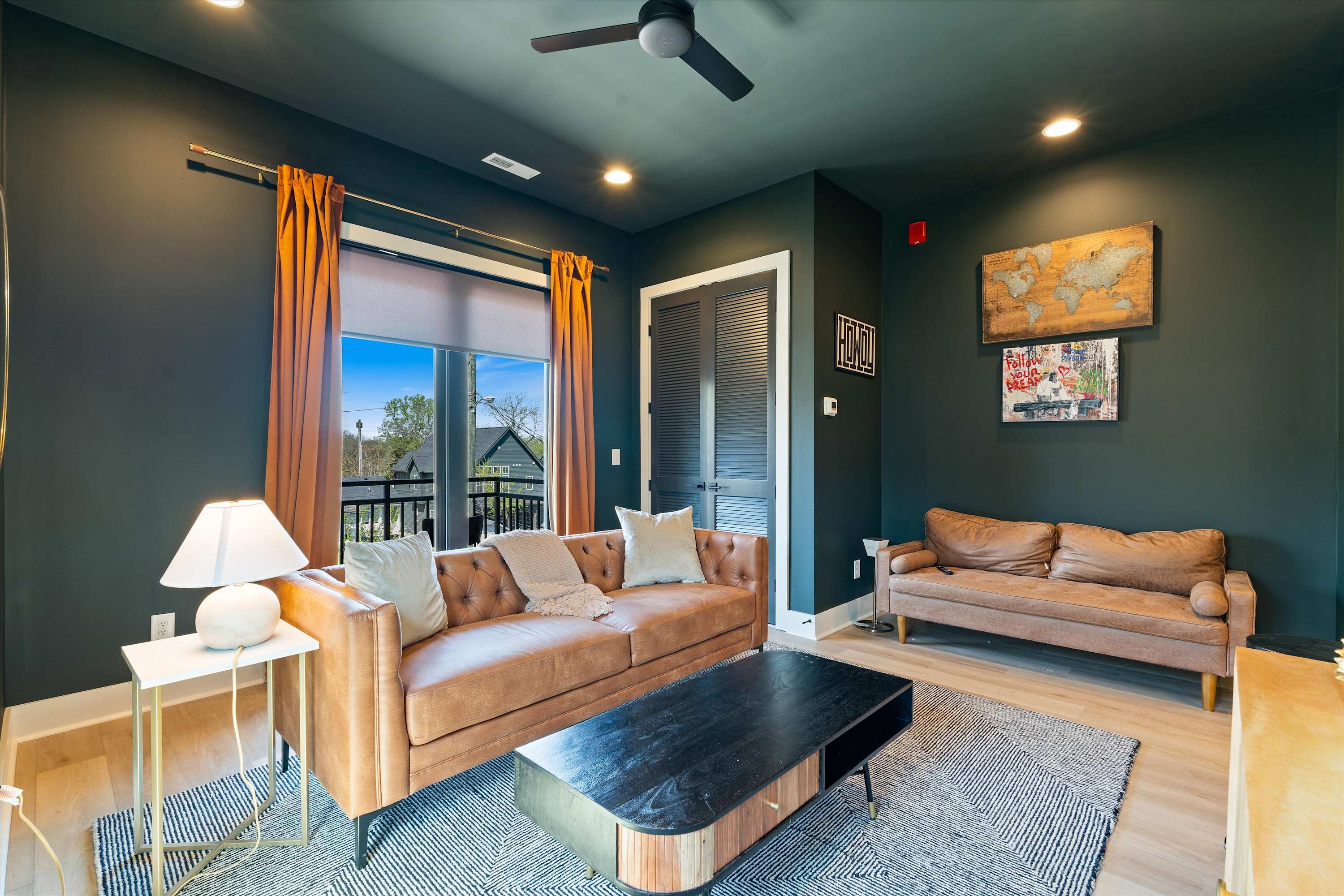 A stylish living room features a brown leather sofa and a matching loveseat, centered around a black and wood coffee table on a geometric area rug. Dark green walls and orange curtains create a bold color scheme, with a large window offering an outdoor view.