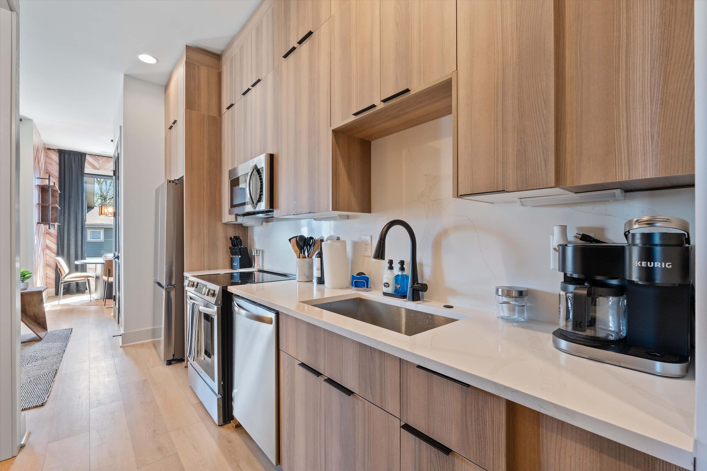 Modern galley kitchen with light wood flat-panel cabinets, white quartz countertops, and stainless steel appliances including a refrigerator, microwave, range, and dishwasher. A black faucet and sink are visible.