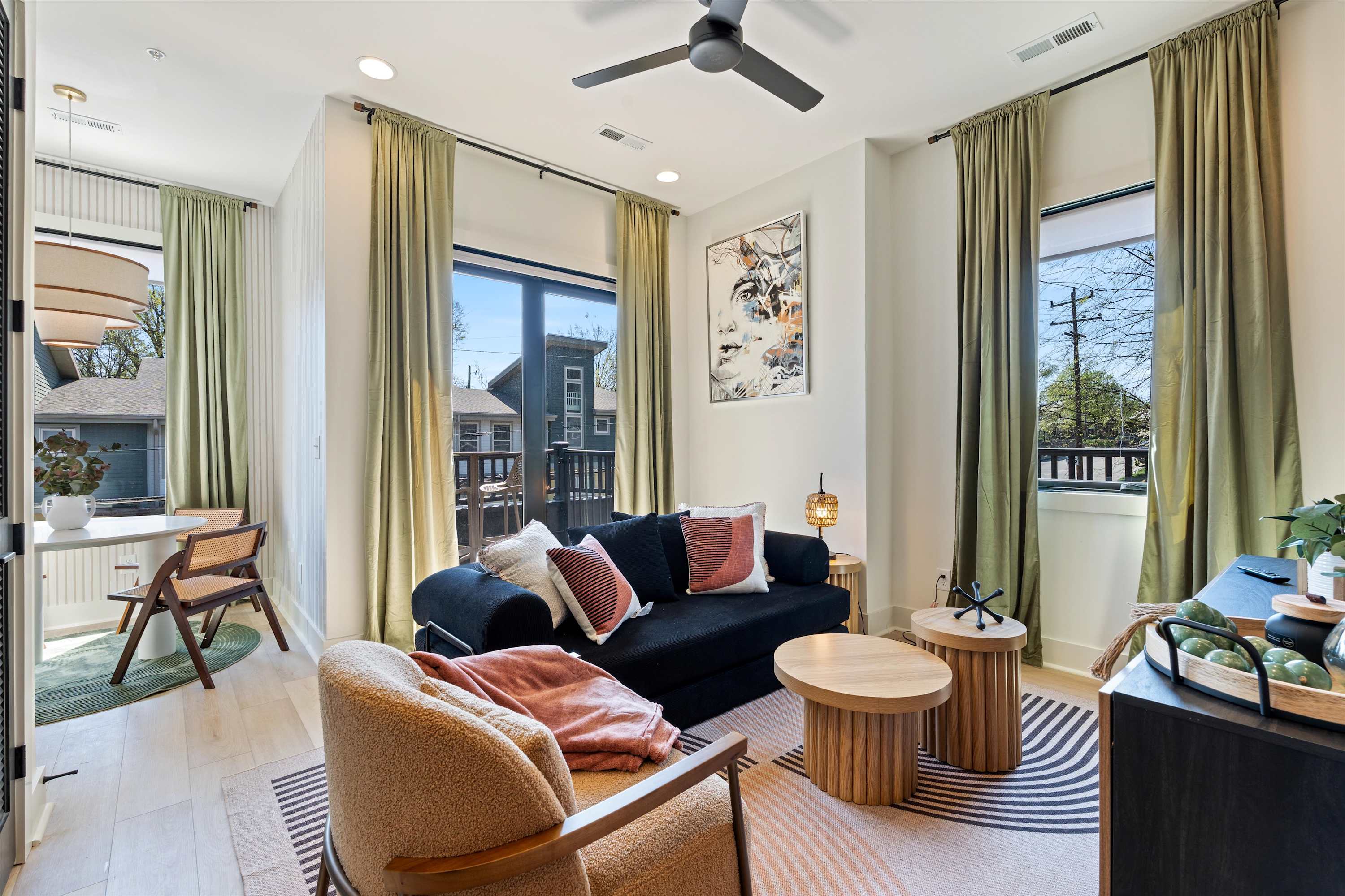 A bright living room with white walls and light wood flooring, featuring a black sofa adorned with patterned pillows and a soft pink throw. Two round, light wood coffee tables sit on a striped rug, while large windows with light green curtains provide natural light and a view of the neighborhood.