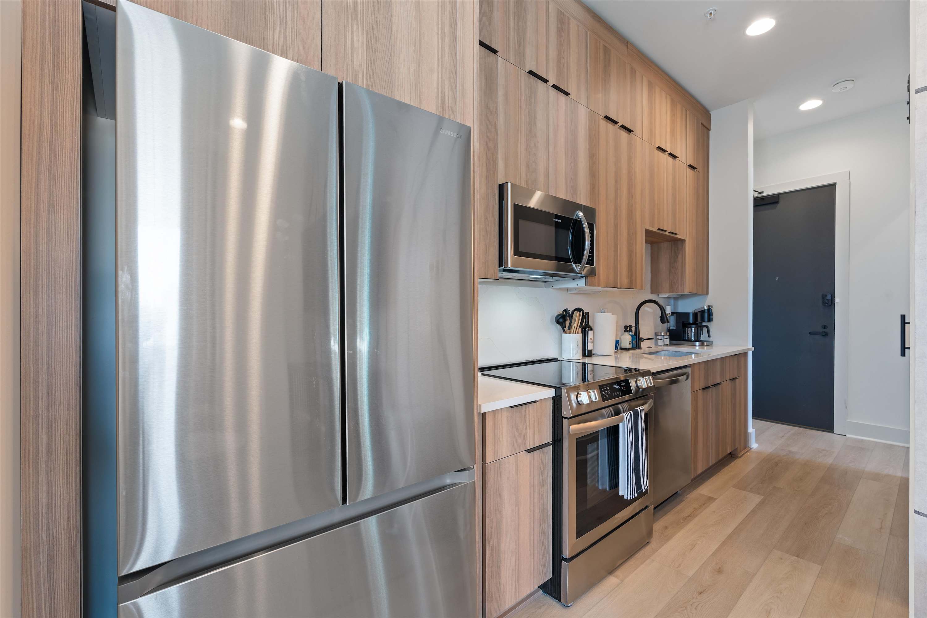 View of the modern kitchen showcasing a large stainless steel French door refrigerator, an electric range, and light wood cabinetry with black hardware. The light wood flooring extends throughout the space.