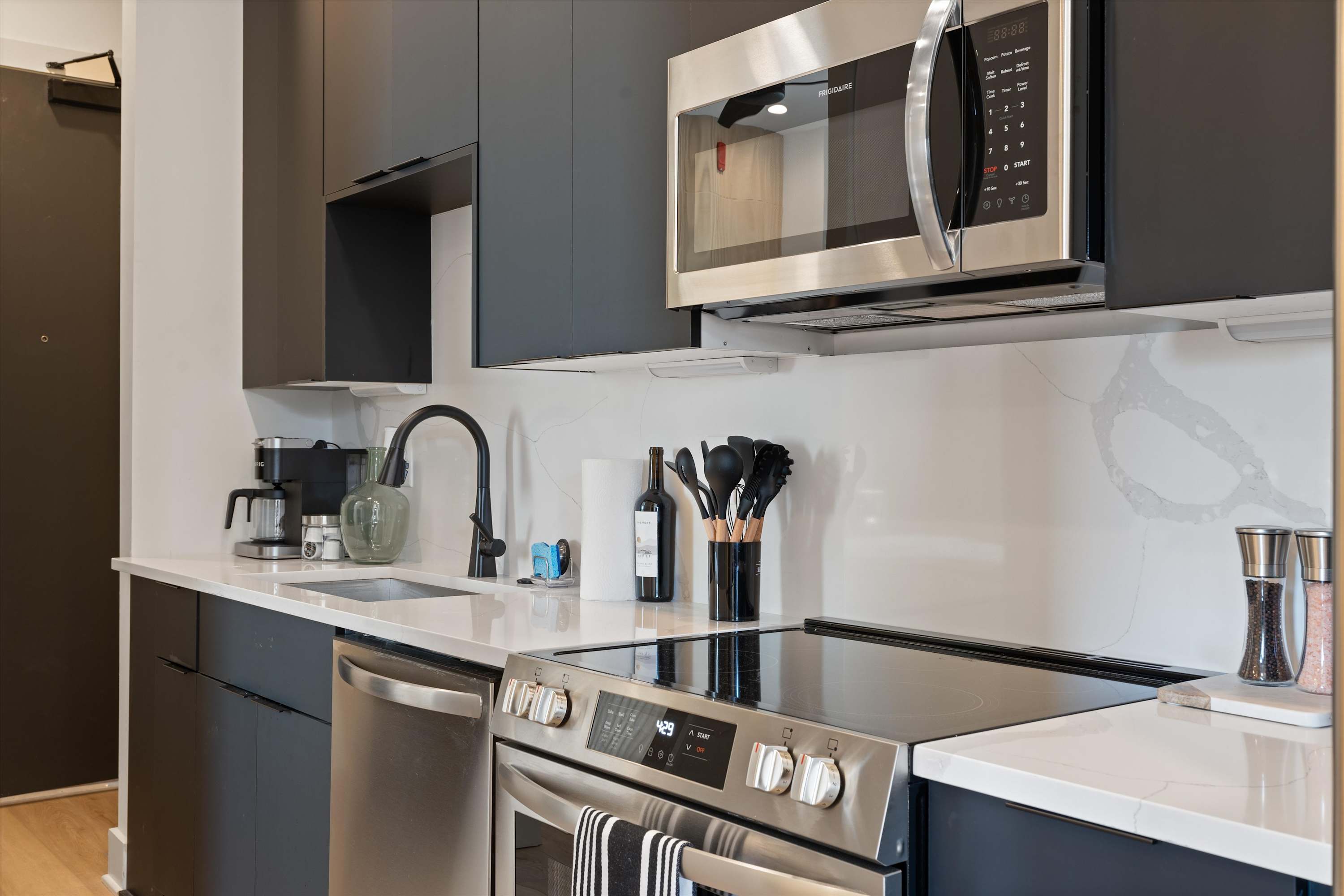 Close-up view of the modern kitchen showing the stainless steel microwave above the electric range, with dark cabinetry and white quartz countertops. Kitchen tools are neatly arranged.