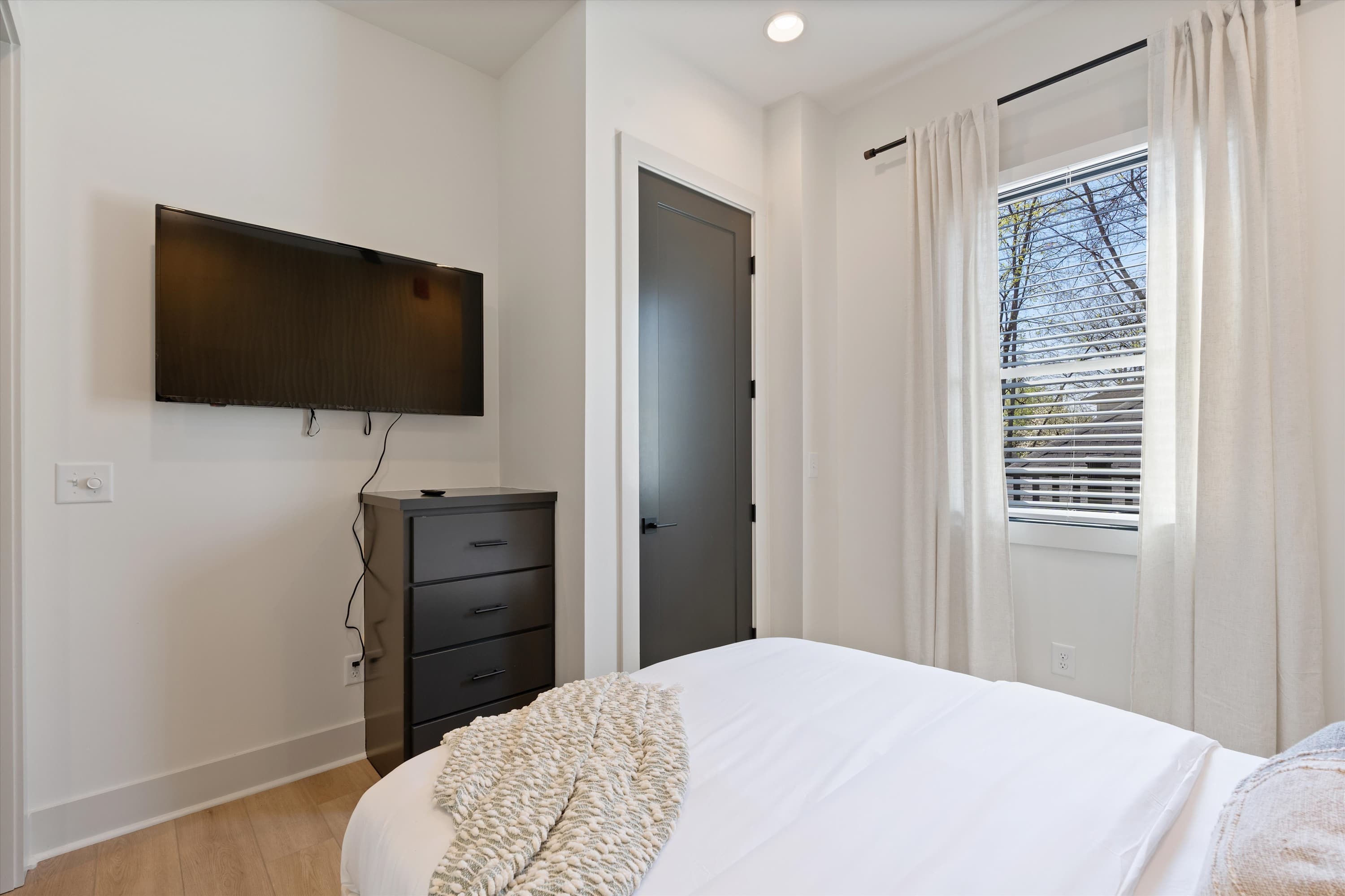A modern bedroom features a white bed with a textured throw, a wall-mounted black flat-screen TV above a matching black three-drawer dresser, and a window with white blinds and sheer curtains.