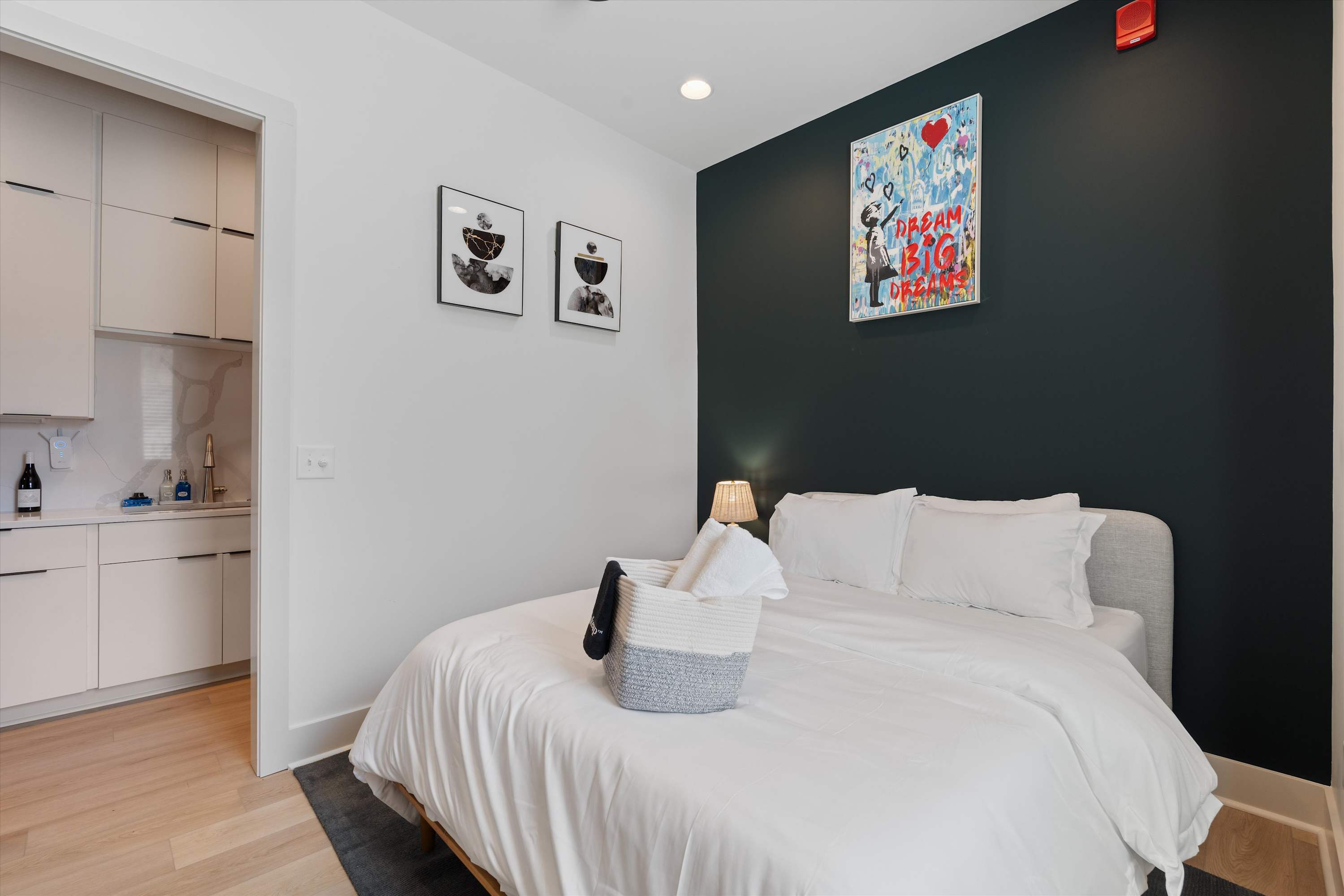 Cozy bedroom with a queen-sized bed featuring white bedding, a dark accent wall behind the headboard, light wood-look flooring, and modern art prints, with a partial view of the kitchen through an open doorway.