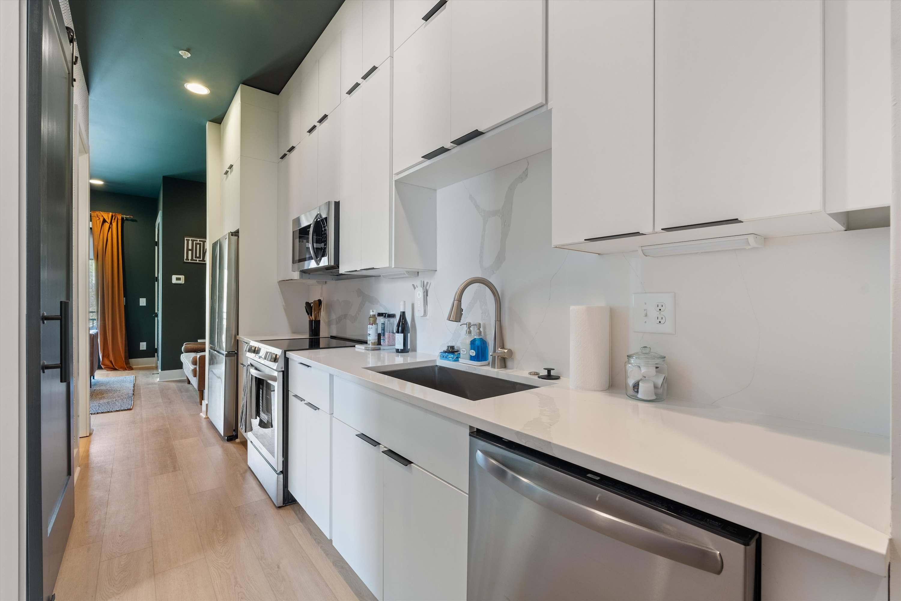 A full view of the modern galley kitchen with white flat-panel cabinets, stainless steel appliances, and light hardwood floors. A dark teal accent wall is visible in the adjacent living area.