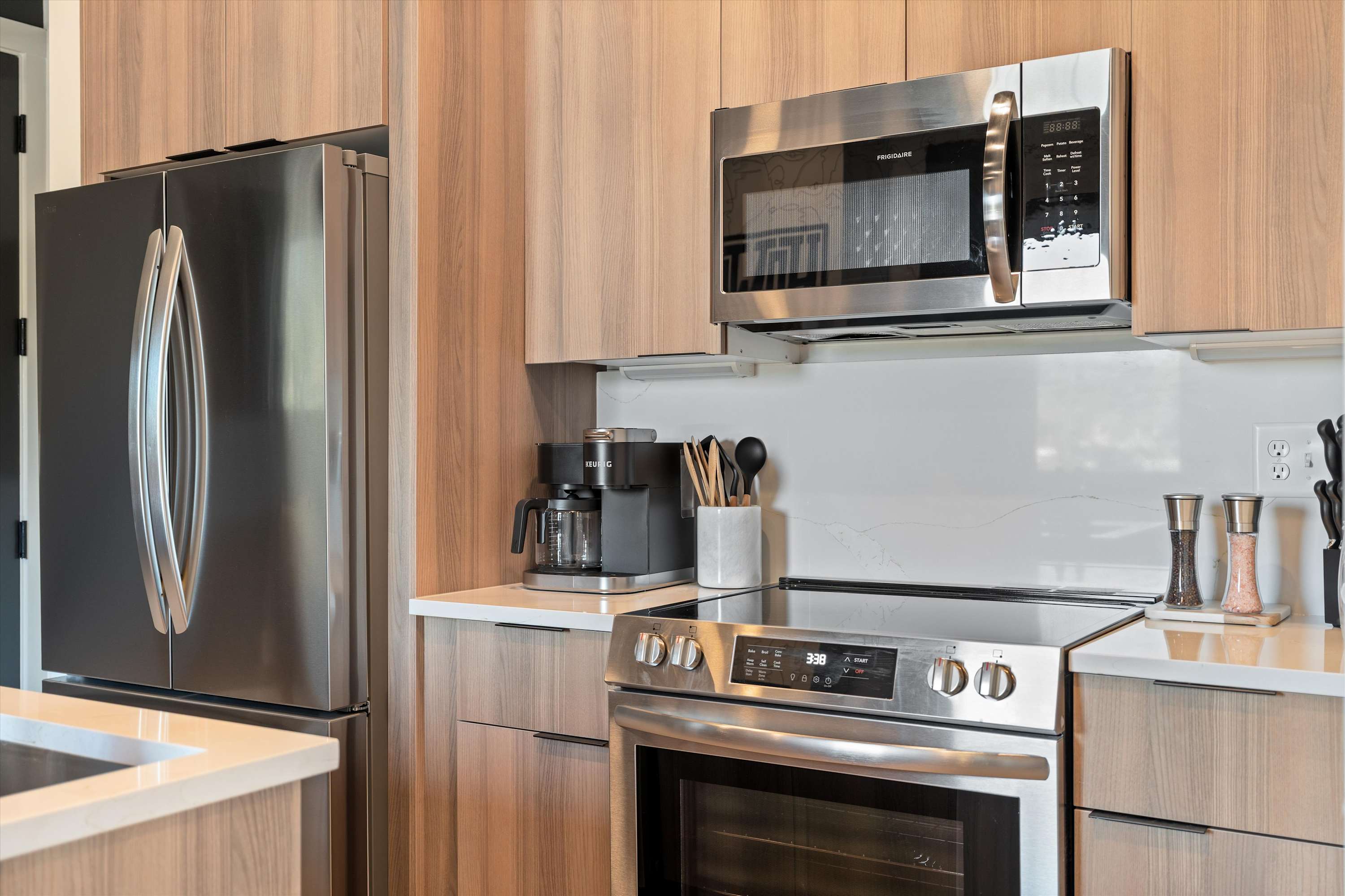 Modern kitchen featuring stainless steel appliances including a refrigerator, microwave, and electric range, set against light wood cabinetry and white quartz countertops.
