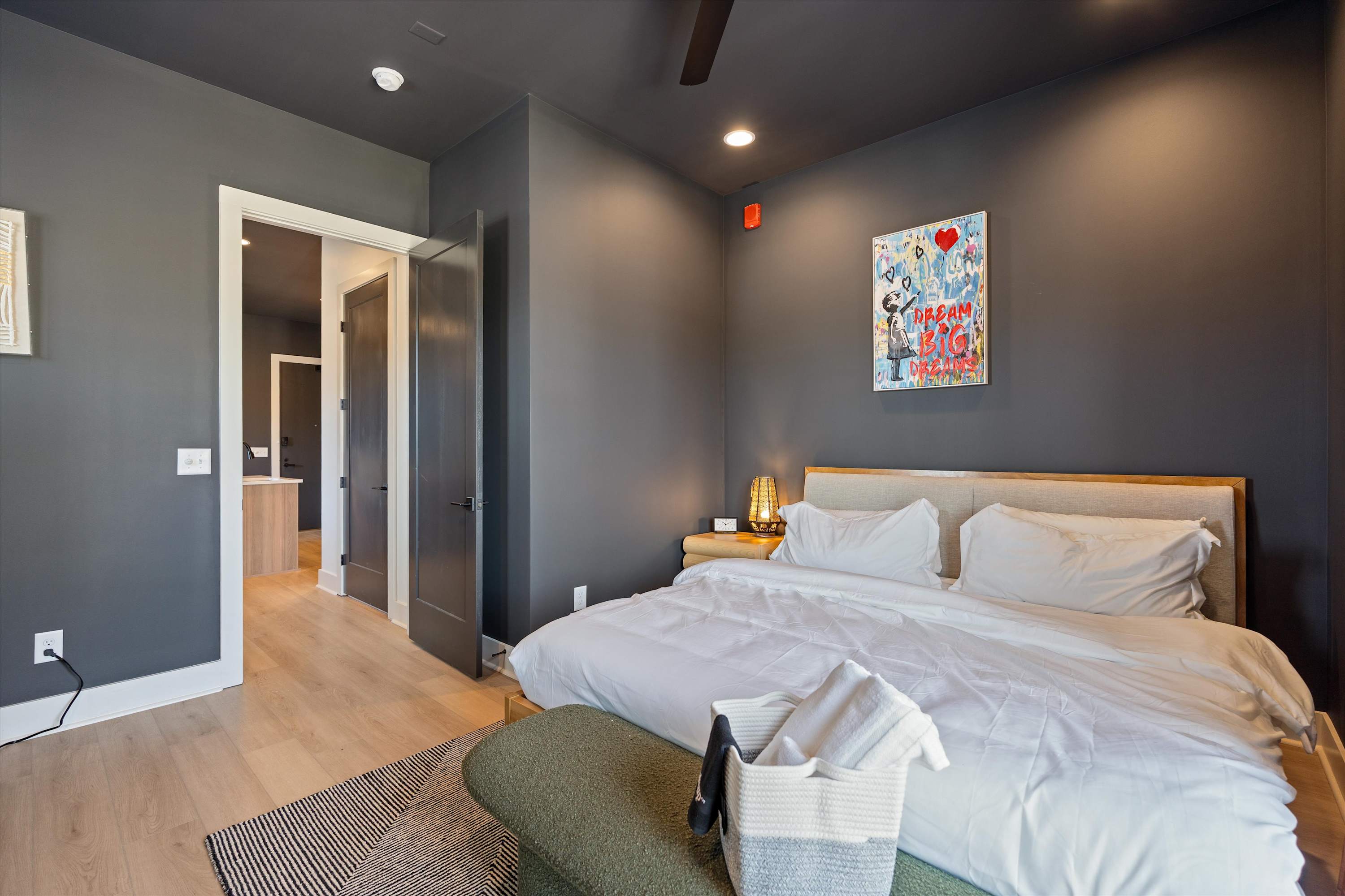 Contemporary bedroom with a king-sized bed, light wood headboard, dark grey walls, and a colorful abstract artwork above the bed, featuring light wood flooring.