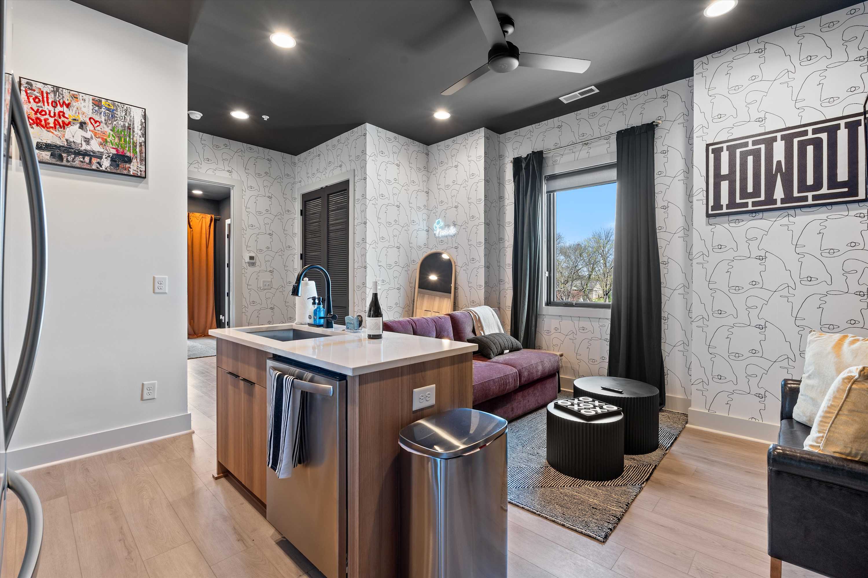 Stylish living area with a plum-colored sofa, two round black coffee tables, and a patterned accent wall featuring abstract faces. A large window with dark curtains offers a view outside, and a full-length mirror leans against the wall.